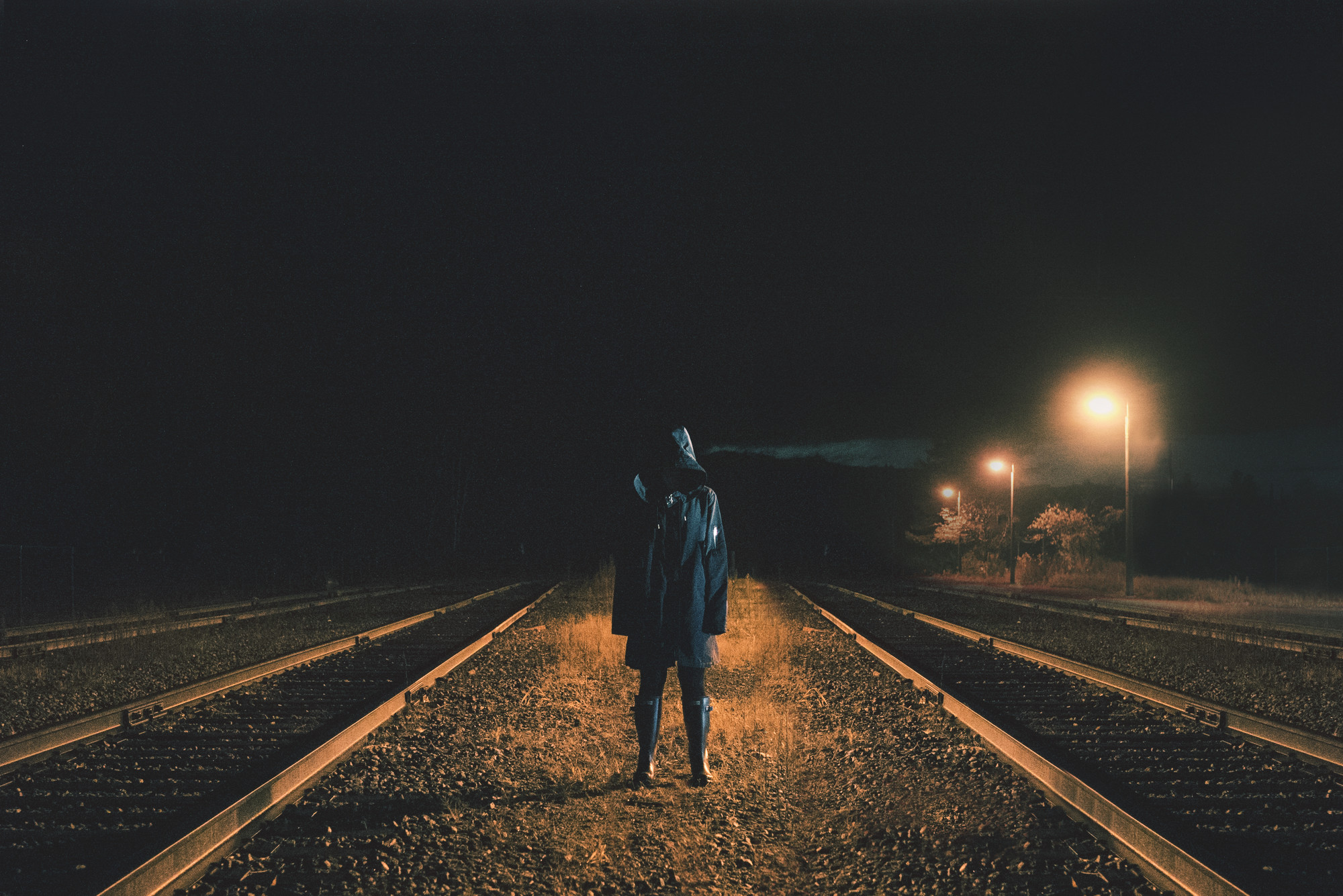 A hooded figure stands on train tracks at night illuminated by streetlights