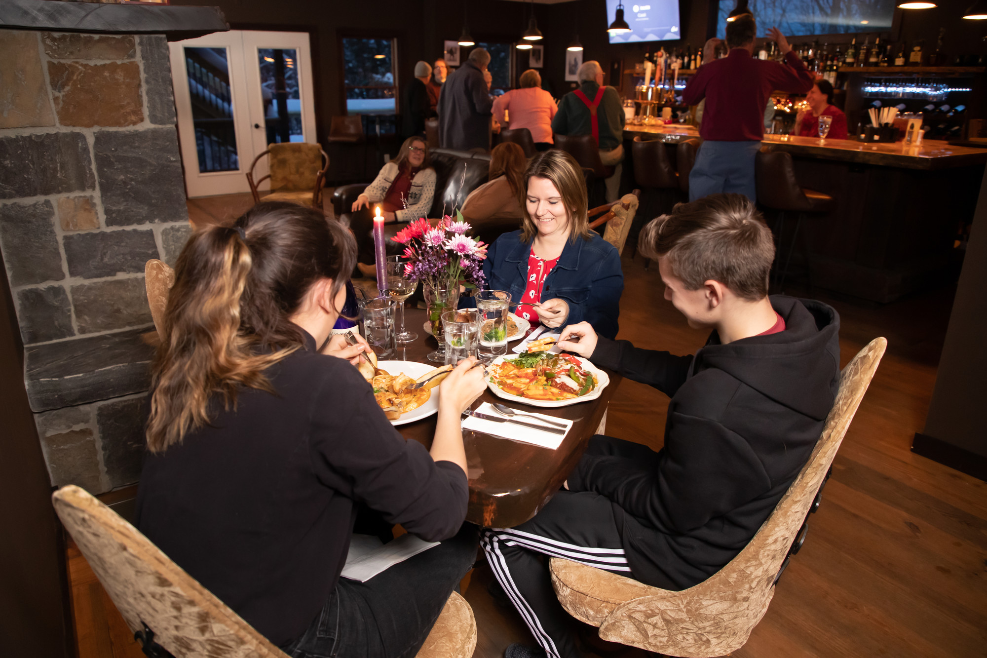 A woman and two teenagers sit at a table, eating, at a lively restaurant.