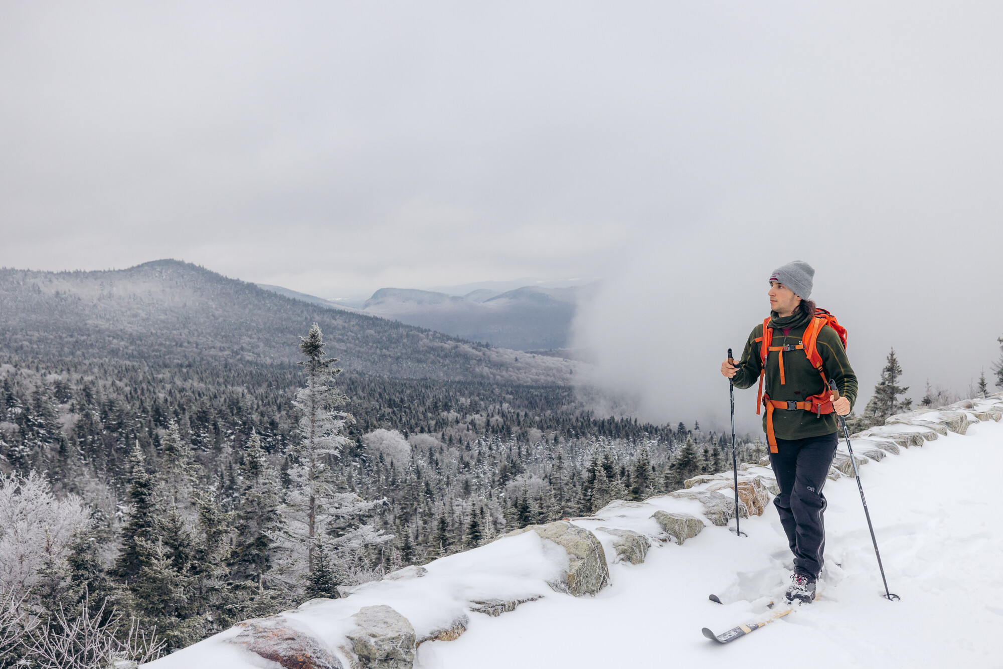 A man cross-country skis up a mountain trail with a view of trees down below. 