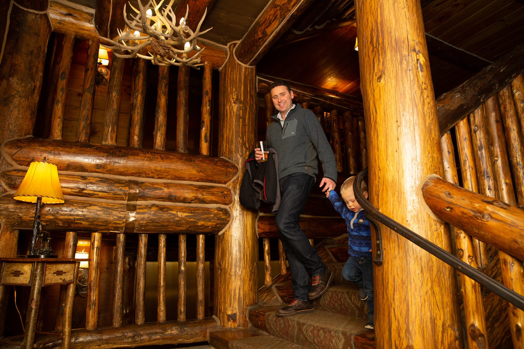 A man and toddler walk down grand log stairs in a lodge.