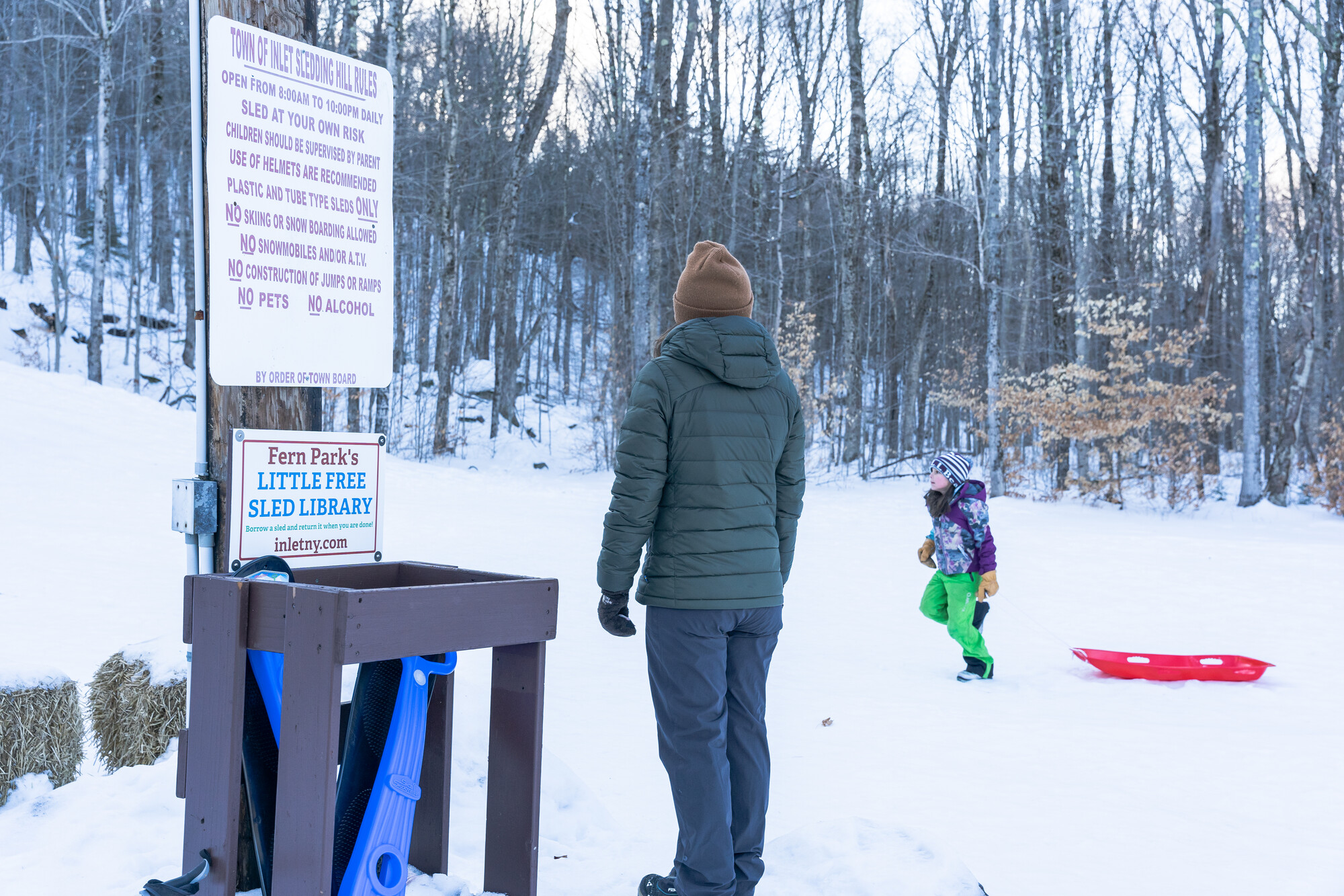 A woman stands next to a "library" of free sleds to borrow, while watching a child on a sled.