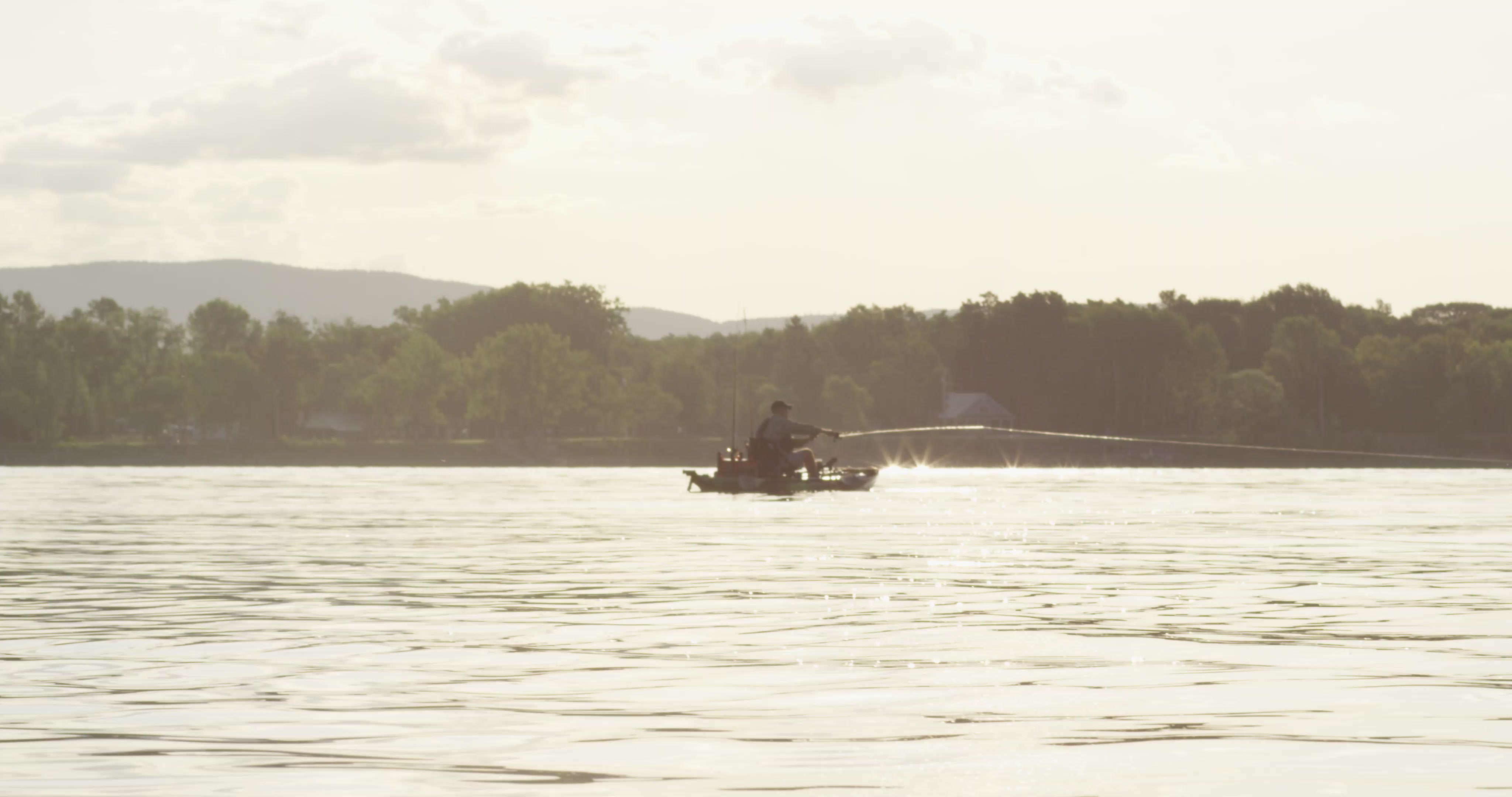 Man stands on kayak, fishing near the Lake Champlain bridge.