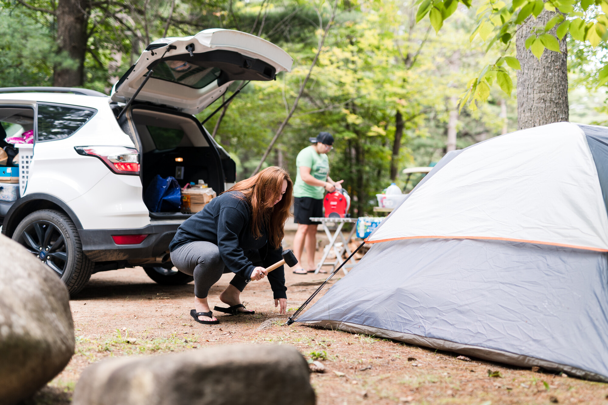 A woman uses a mallet to pound a tent stake at a campground surrounded by tres.