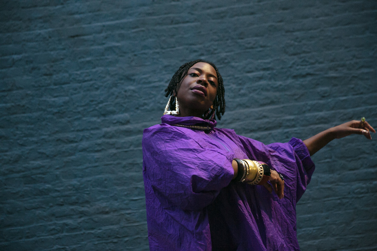 A black woman with braided hair, wearing a bright purple dress poses in front of a blue brick wall.