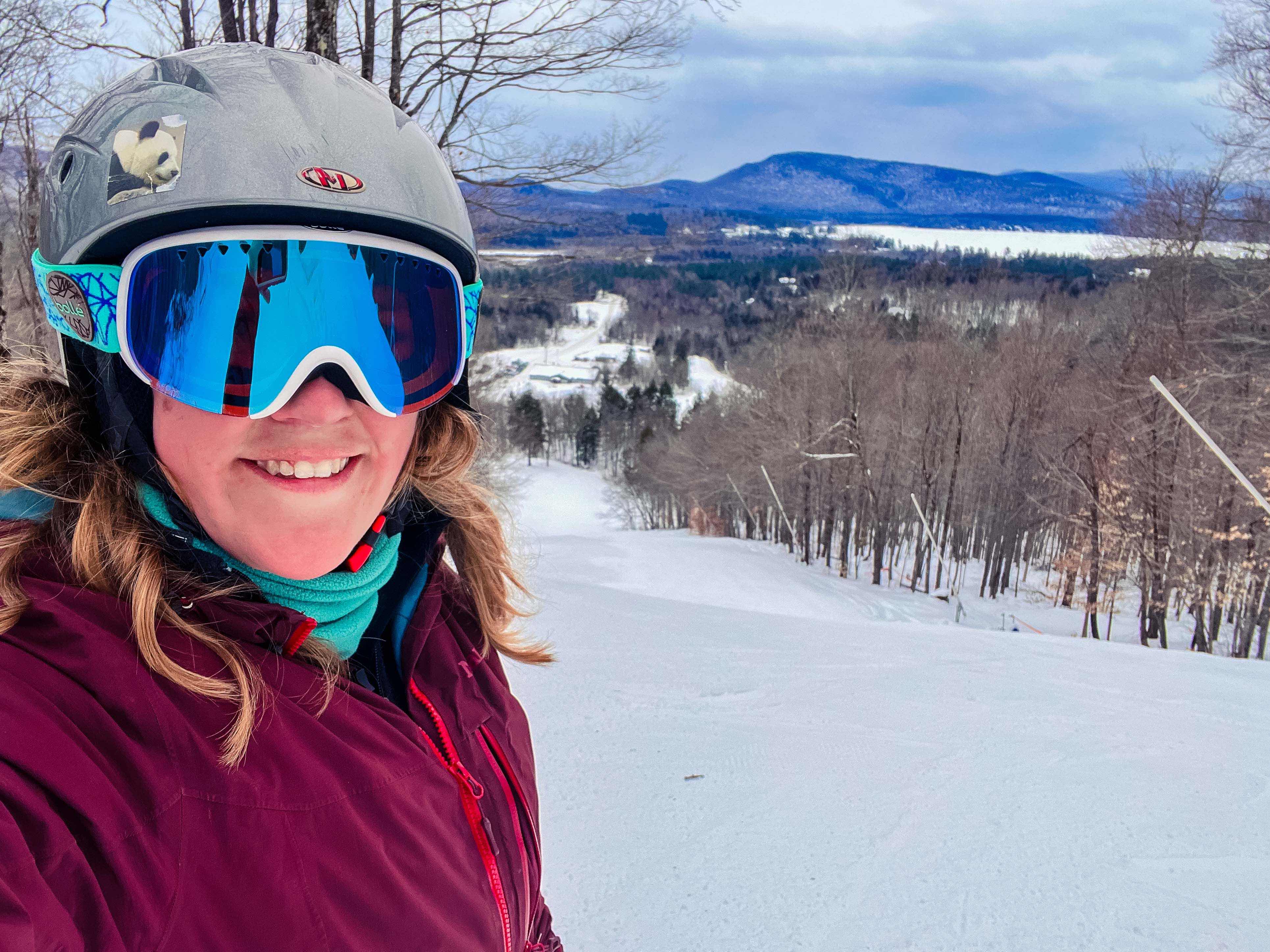 A skier with goggles and a helmet and purple coat smiling for a selfie at the top of a ski run. 