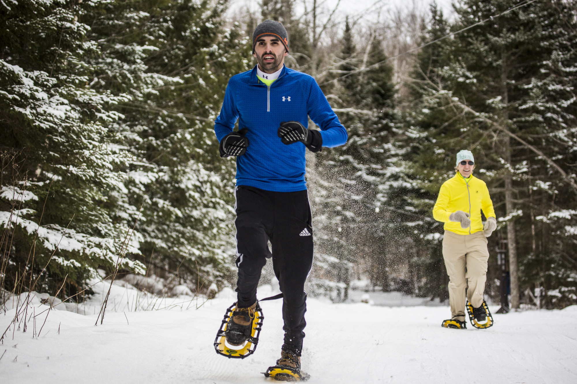 A man and woman run in snowshoes on a snowy trail. 