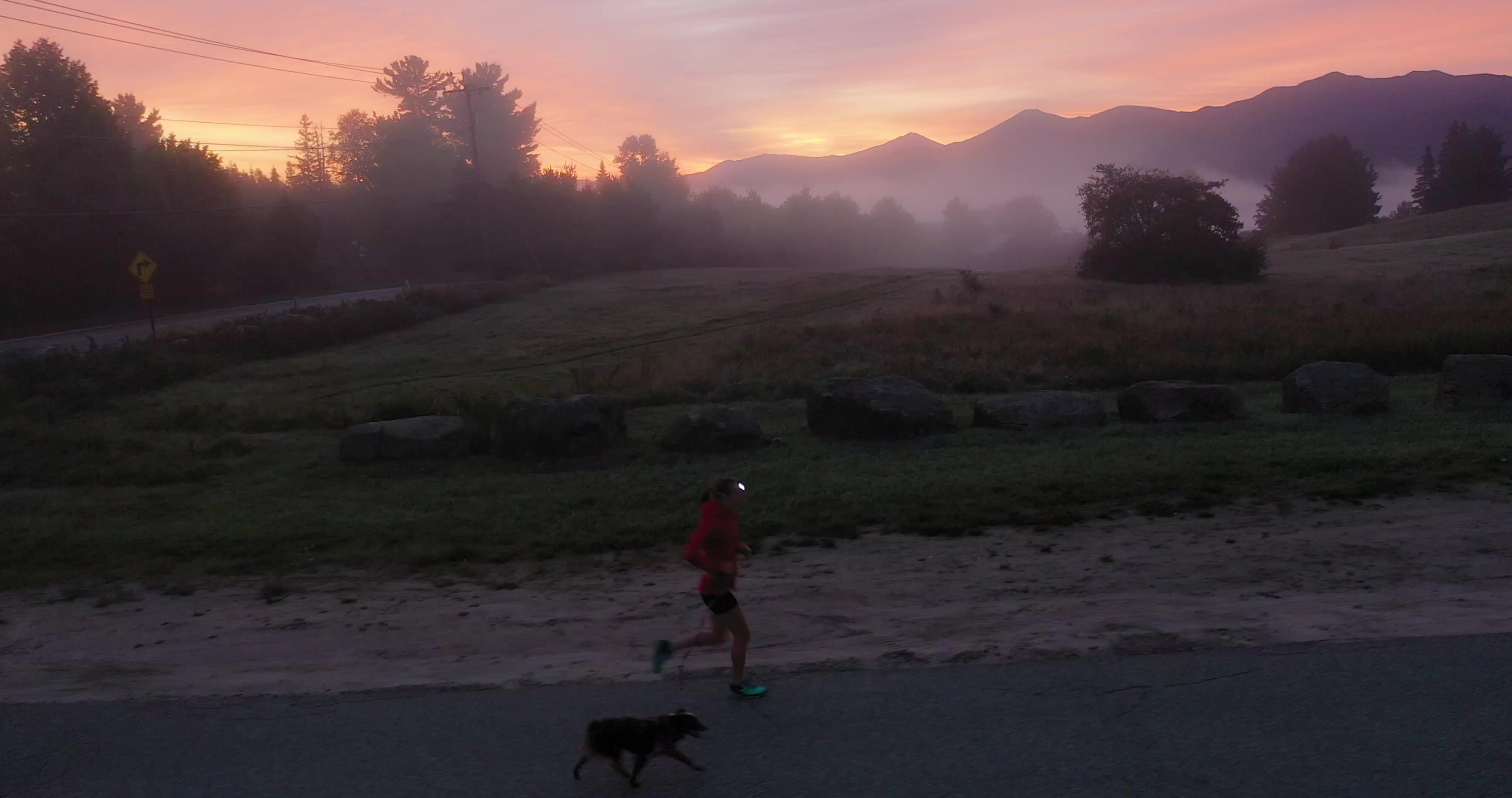 An athlete runs up a road with her dog as the sun rises among the mountains behind them