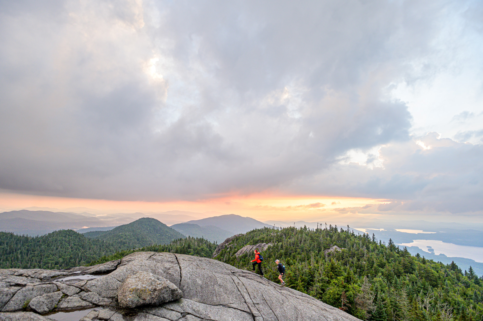 Two hikers climb on summit rocks as the sun sets with an orange glow.