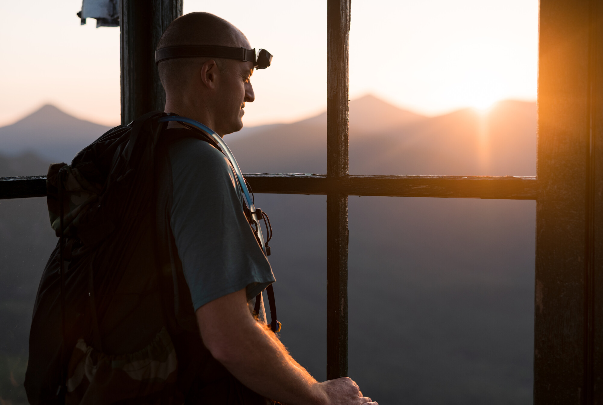 A man takes in the view from a fire tower cab as the sun rises behind the mountains. 