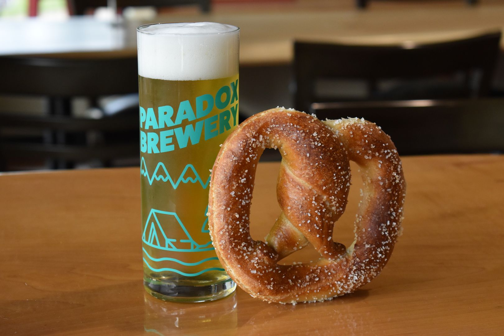 A tall, narrow glass of beer sits on a wooden bar next to a big soft pretzel