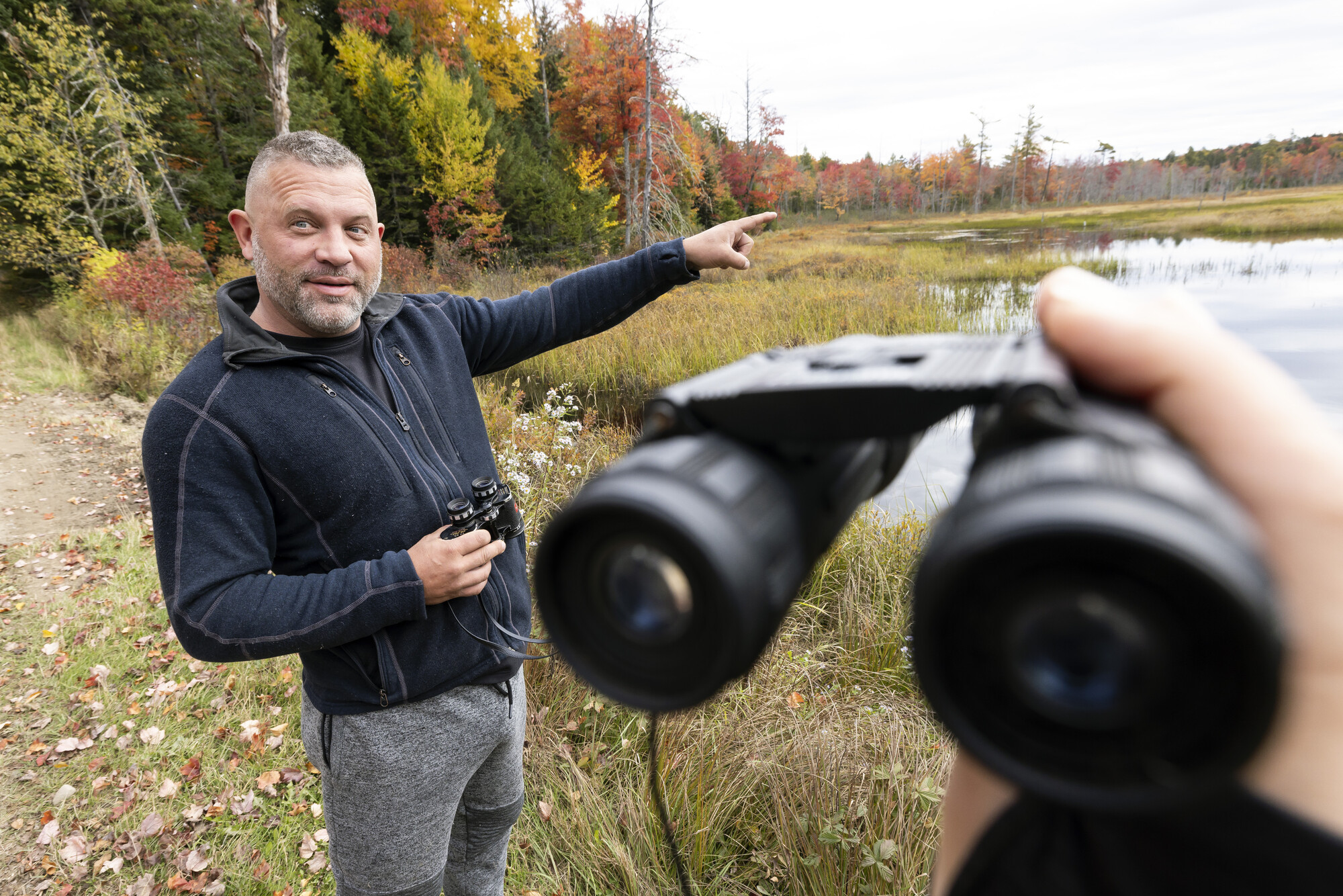 A view of a man pointing to marshland for the viewer to look with binoculars. 