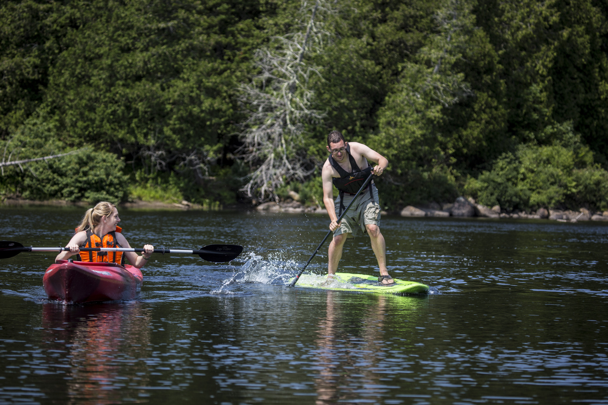 A young lady kayaks alongside a young man paddling on a stand-up paddle board.
