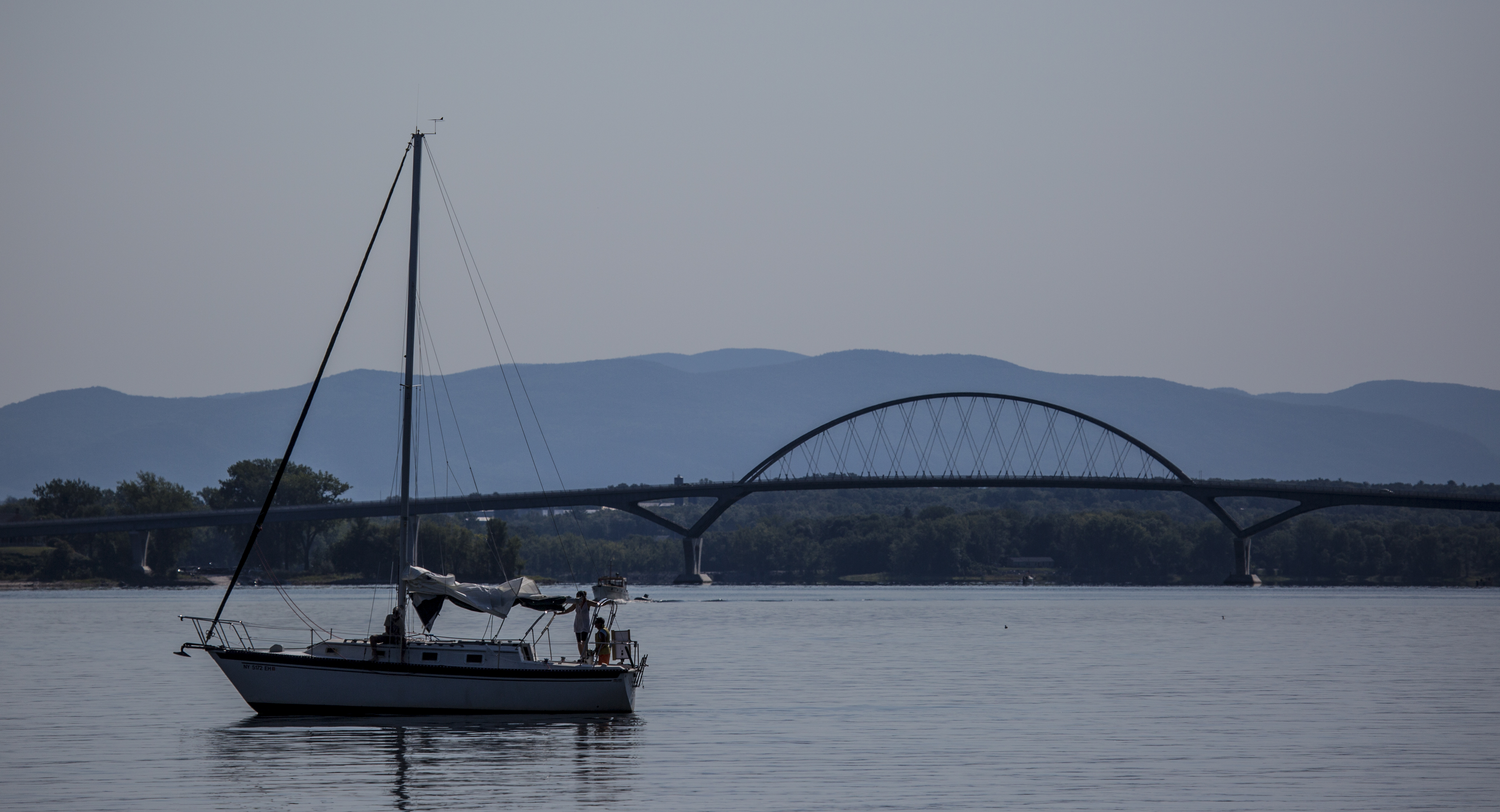A sailboat with two people onboard with a bridge and mountains in the background.