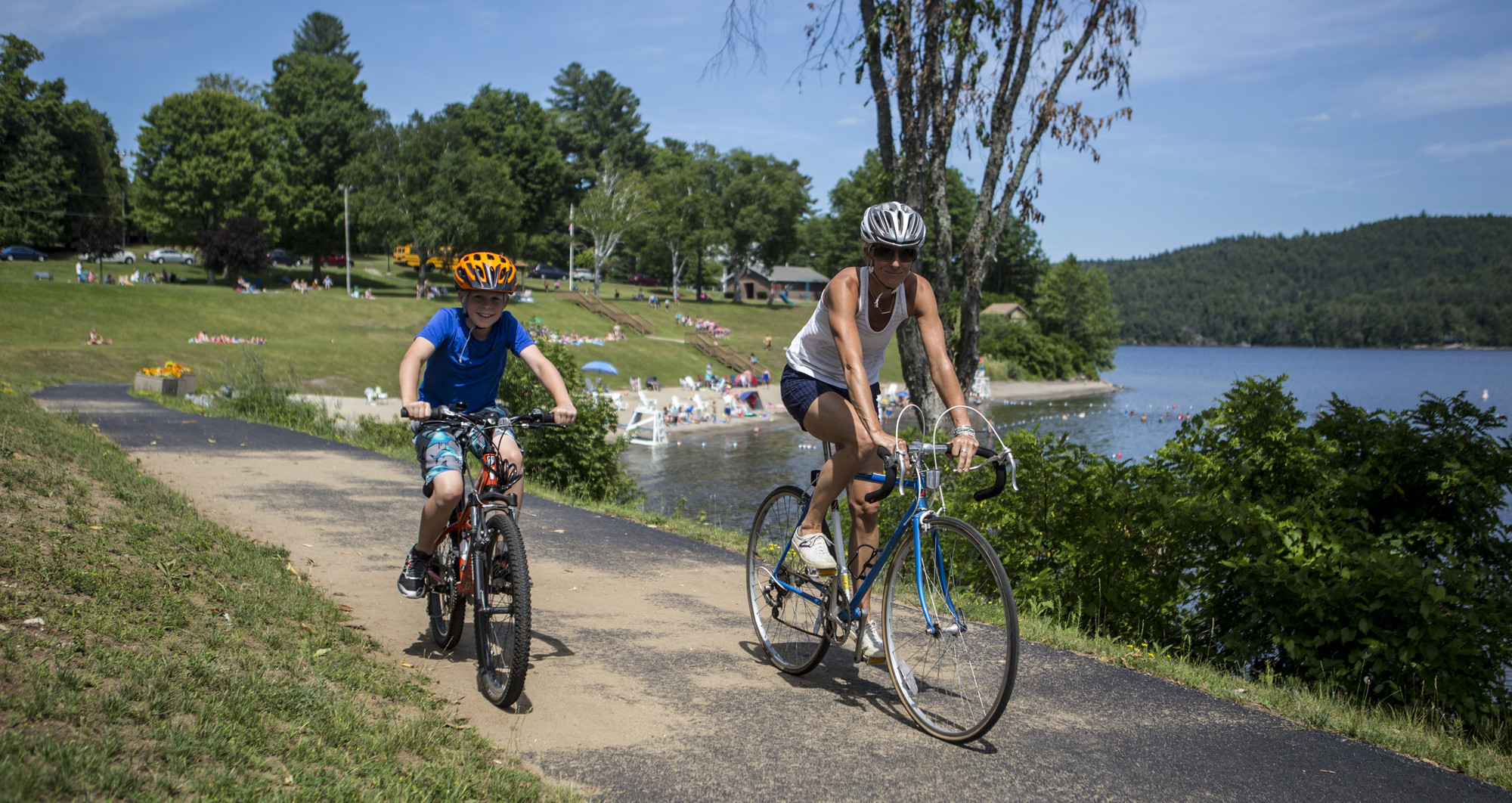 A woman and boy ride bicycles on a path with a large lake in the background.