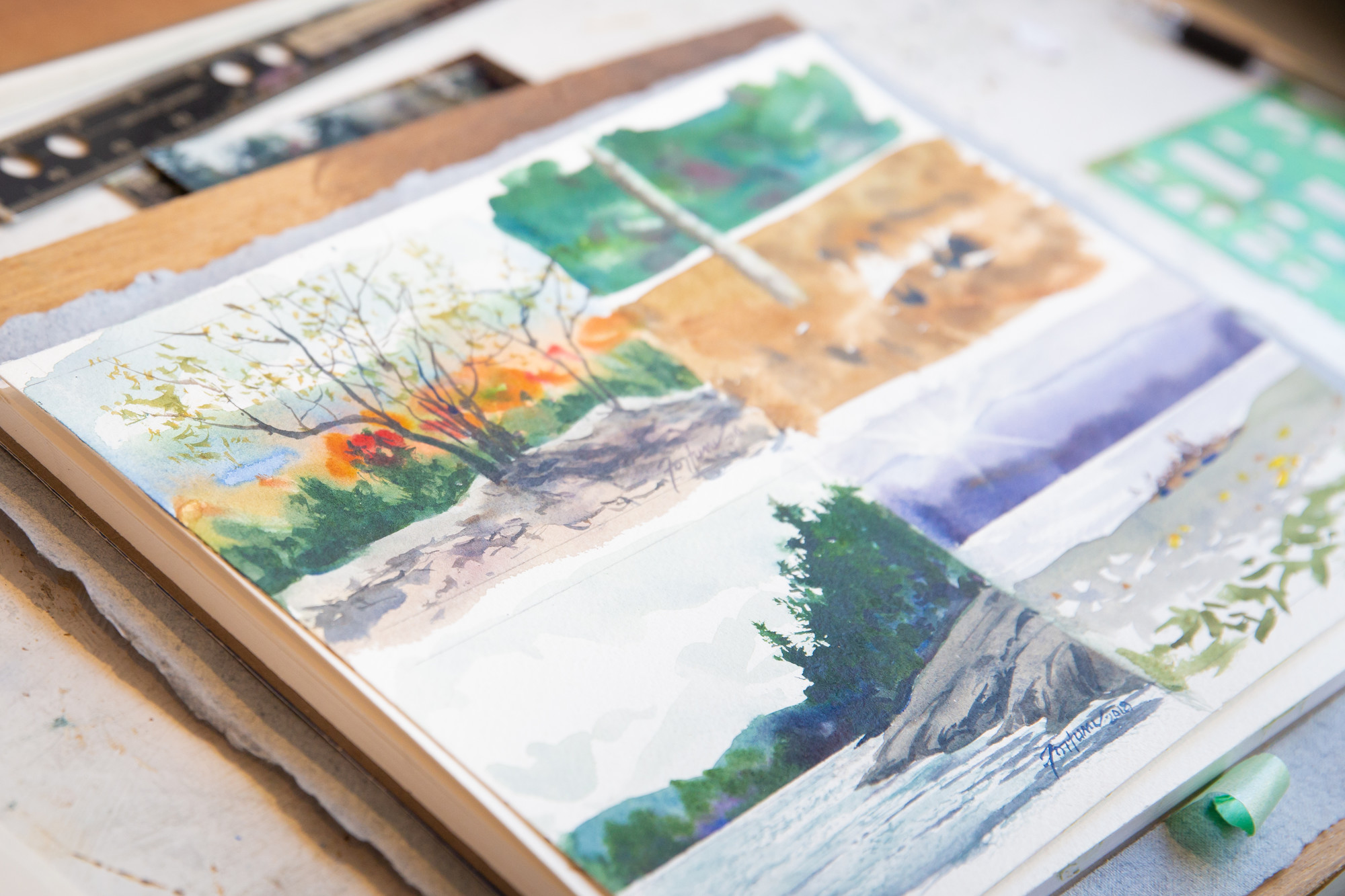 A close-up of vibrant watercolor landscape paintings depicting seasonal Adirondack scenes, displayed on an artist’s workspace.