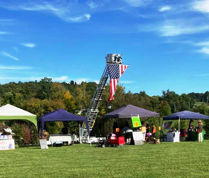 A fire truck and vendor tents at a grassy, tree-lined park on a sunny day.