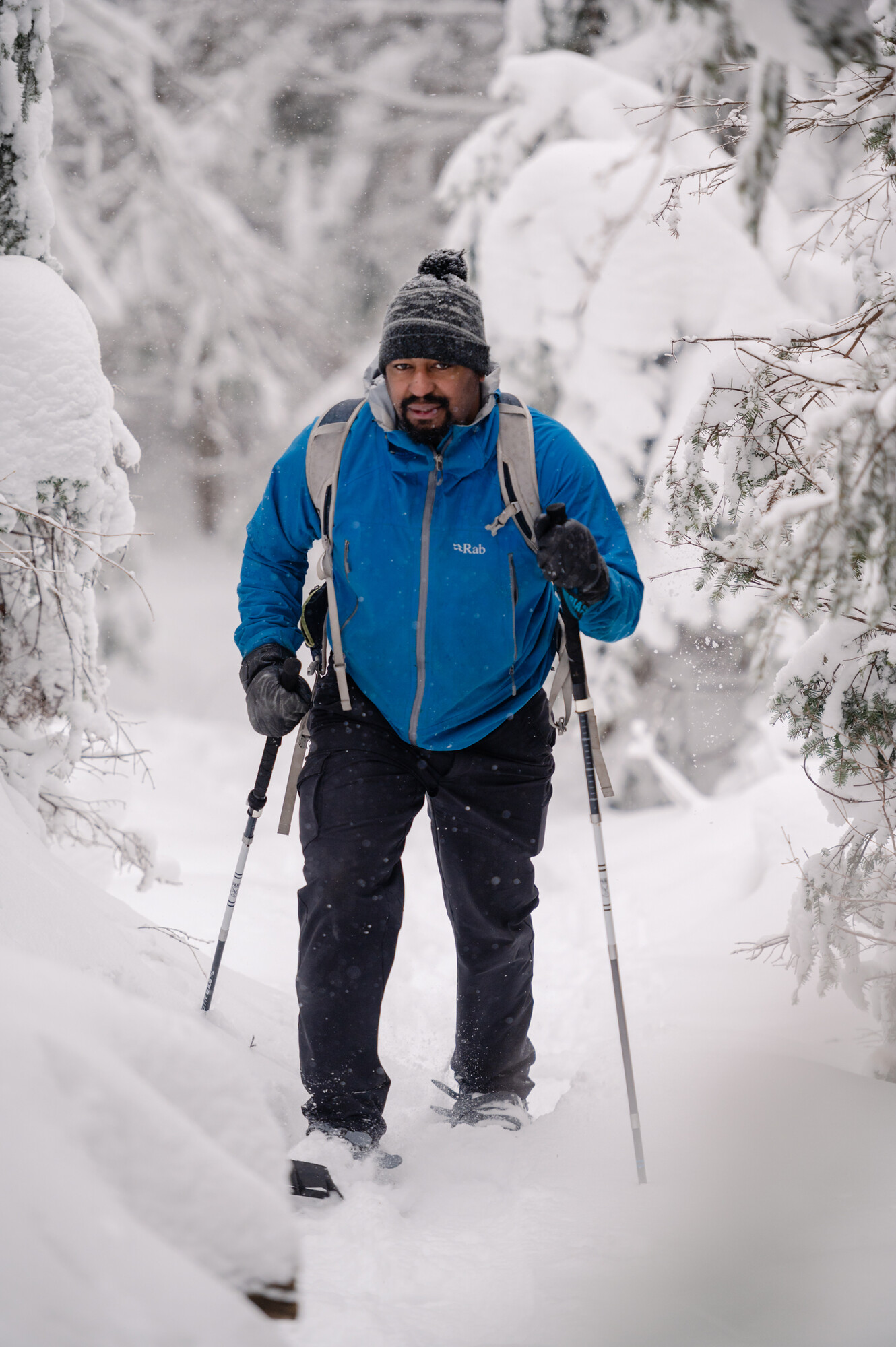 A man in a blue jacket snowshoes as it snows on a winter trail. 