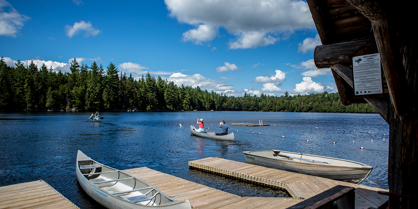 Paddlers in silver canoes depart from a small row of docks on a sparkling blue lake with wooded shoreline in the background.