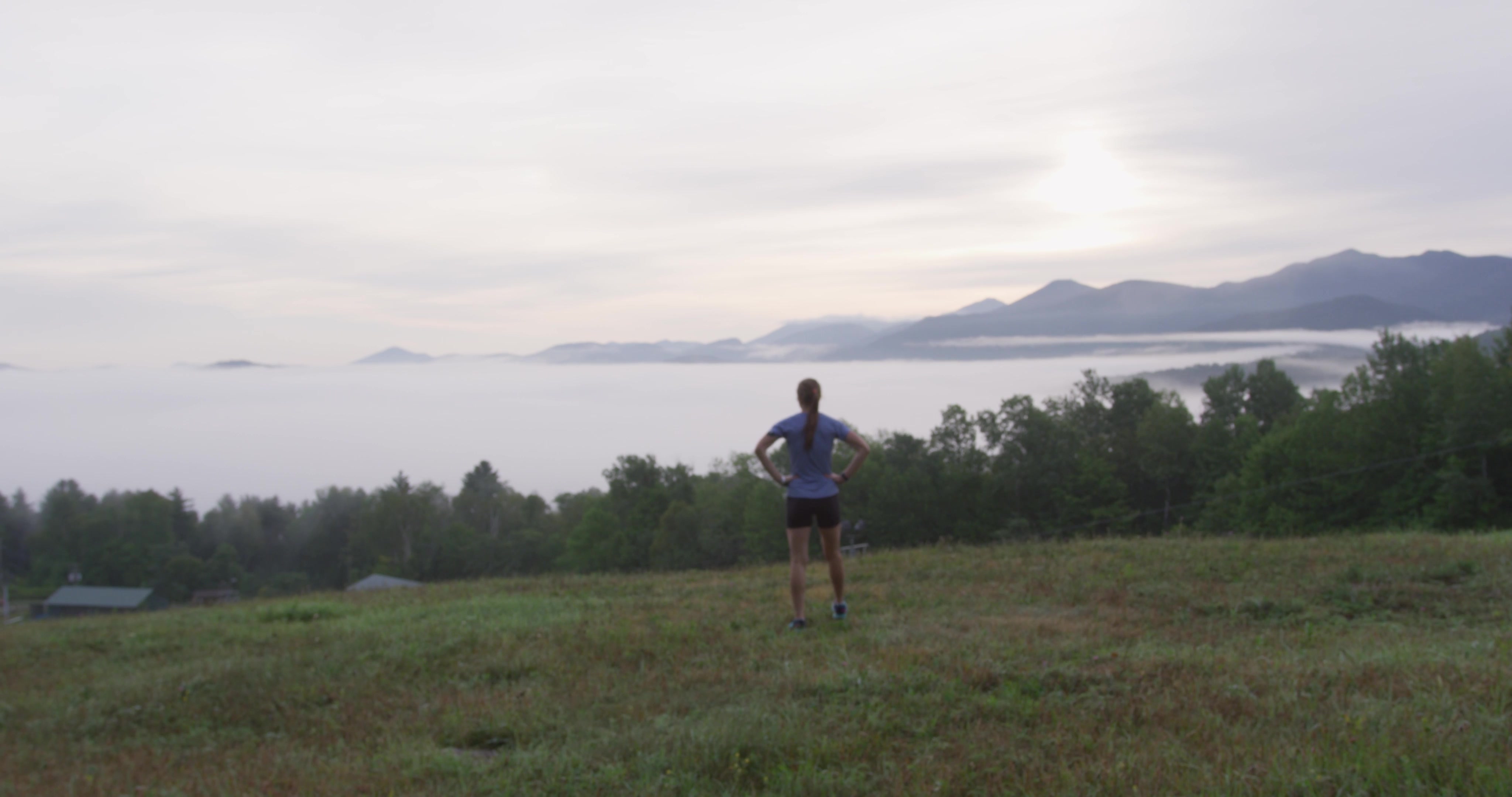 A woman stands at the top of a grassy summit while looking at the mountains along the horizon