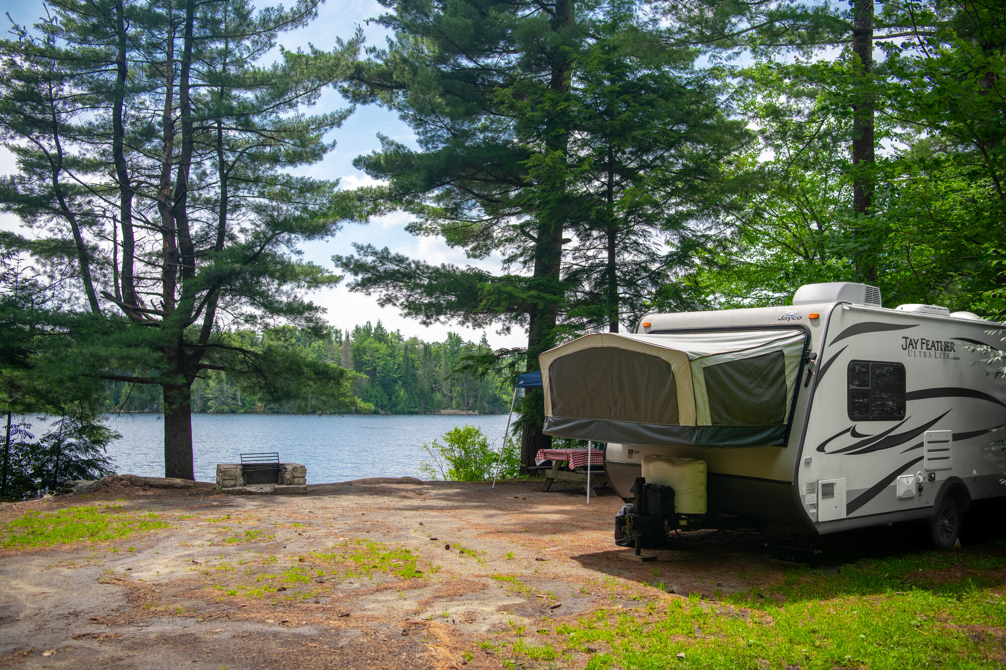 A pop-out camper is opened on the side of a campsite with an empty firepit, overlooking a lake. 