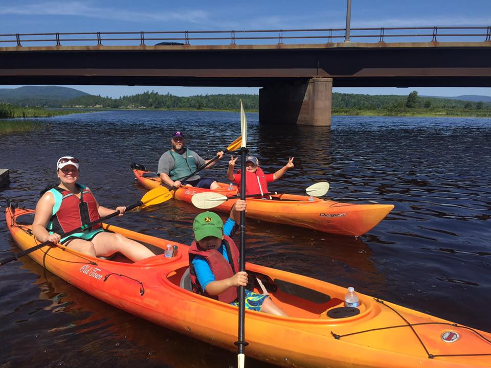 Two kayaks with adults and young children are paddled along a lake.