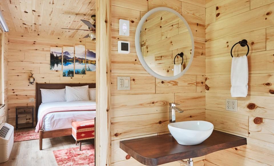 A view of a motel room bathroom and bedroom, showing pale wood paneling, simple but elegant decor, and bright natural lighting.