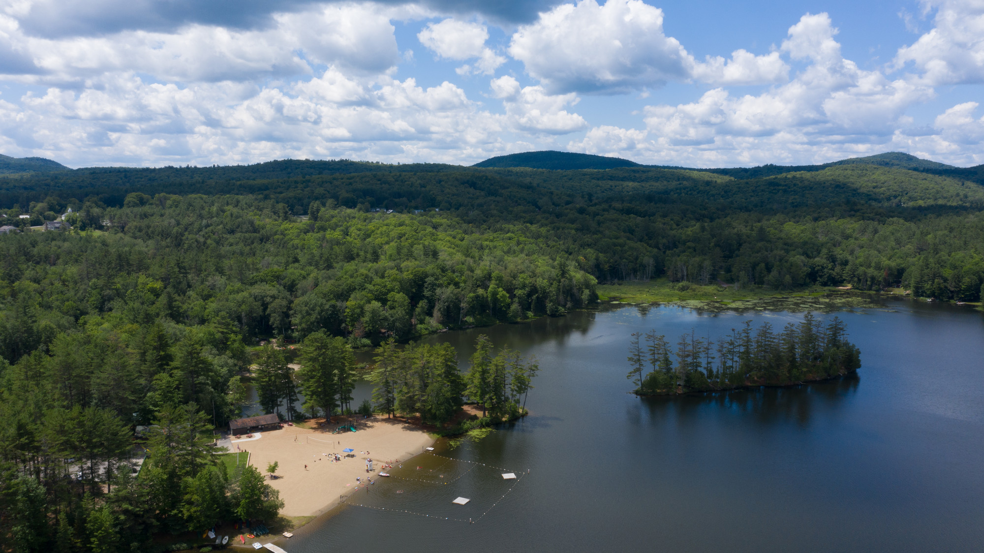 An aerial view of an Adirondack lake and sandy beach, surrounded by thick forests.