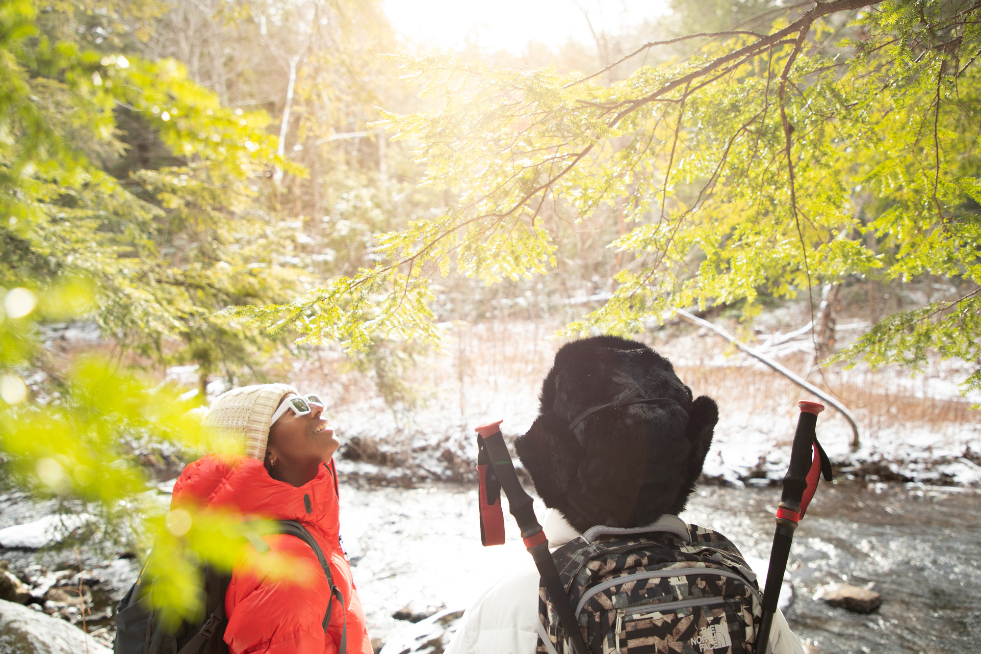 Two people in coats and hats walk through a trail. Water runs behind them. 