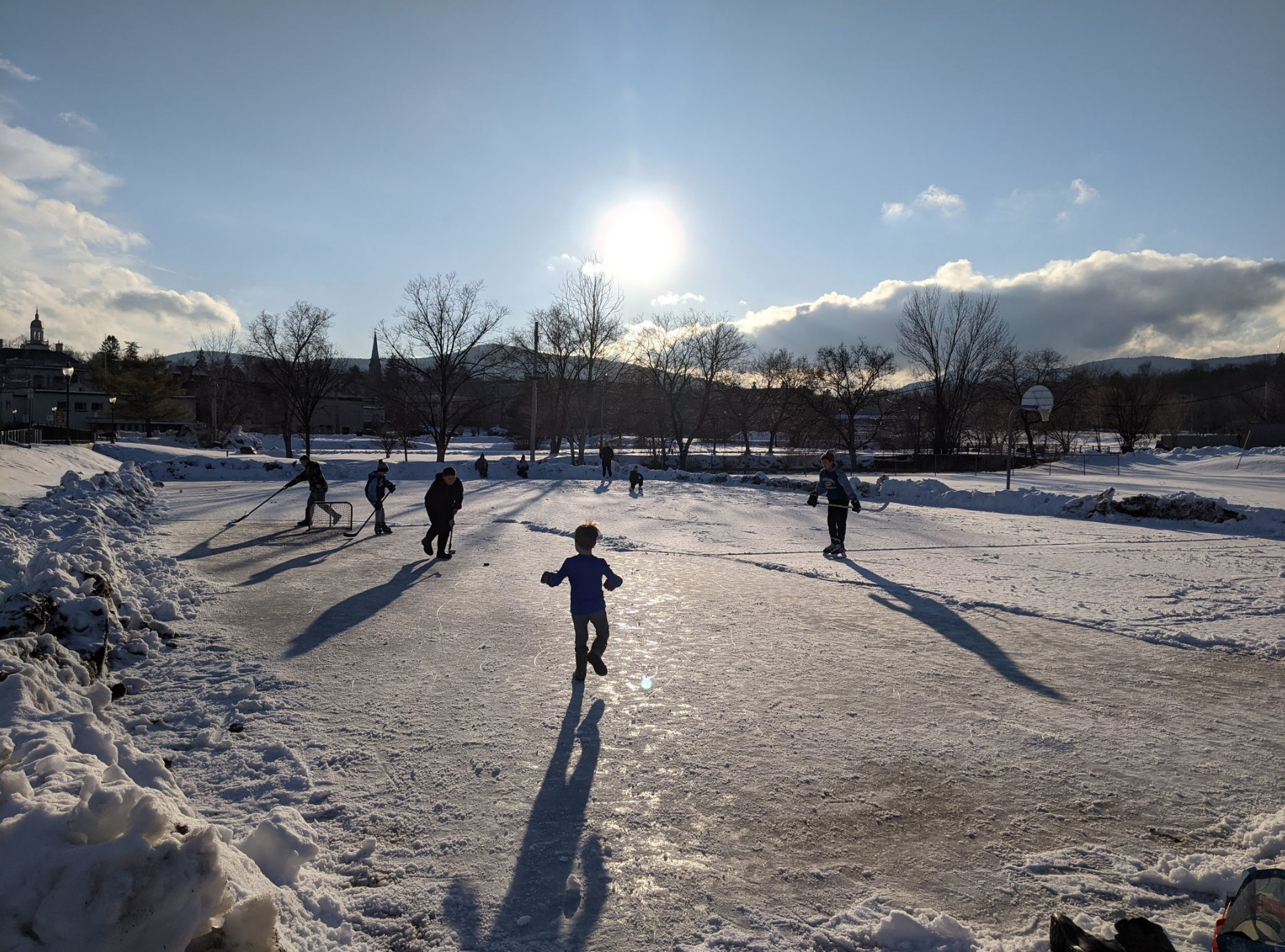 Children and adults play hockey on a large ice skating rink.
