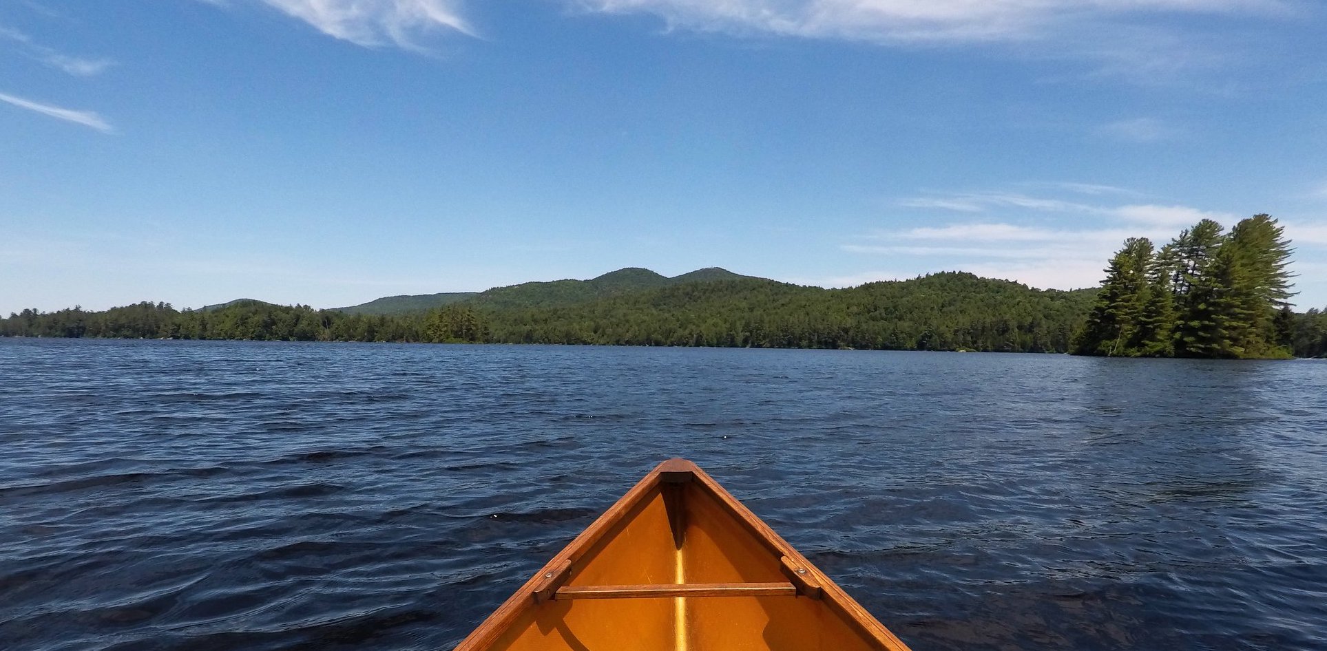 St. Regis Pond The bow of a canoe with a big mountain on the far shoreline and trees with green leaves.