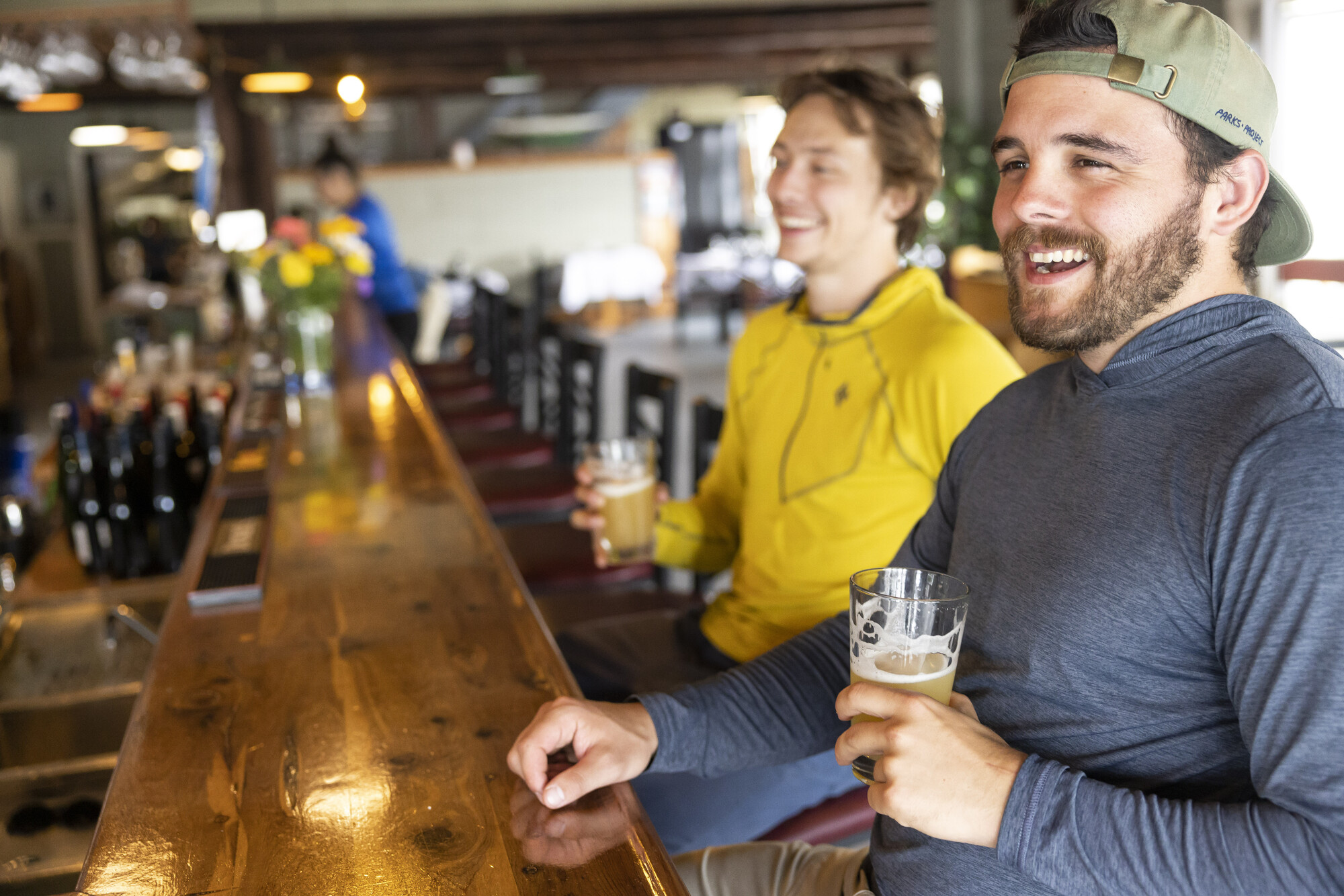 Two men sit at a wooden bar, smiling with golden-colored beer in glasses.