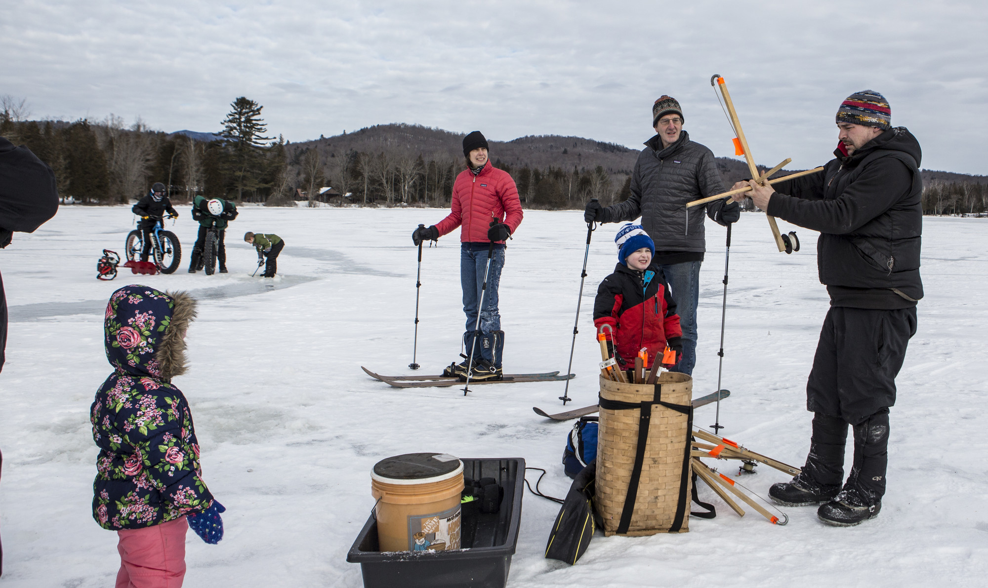 A group of people and children on a lake participating in ice fishing, cross-country skiing, and fat tire biking.