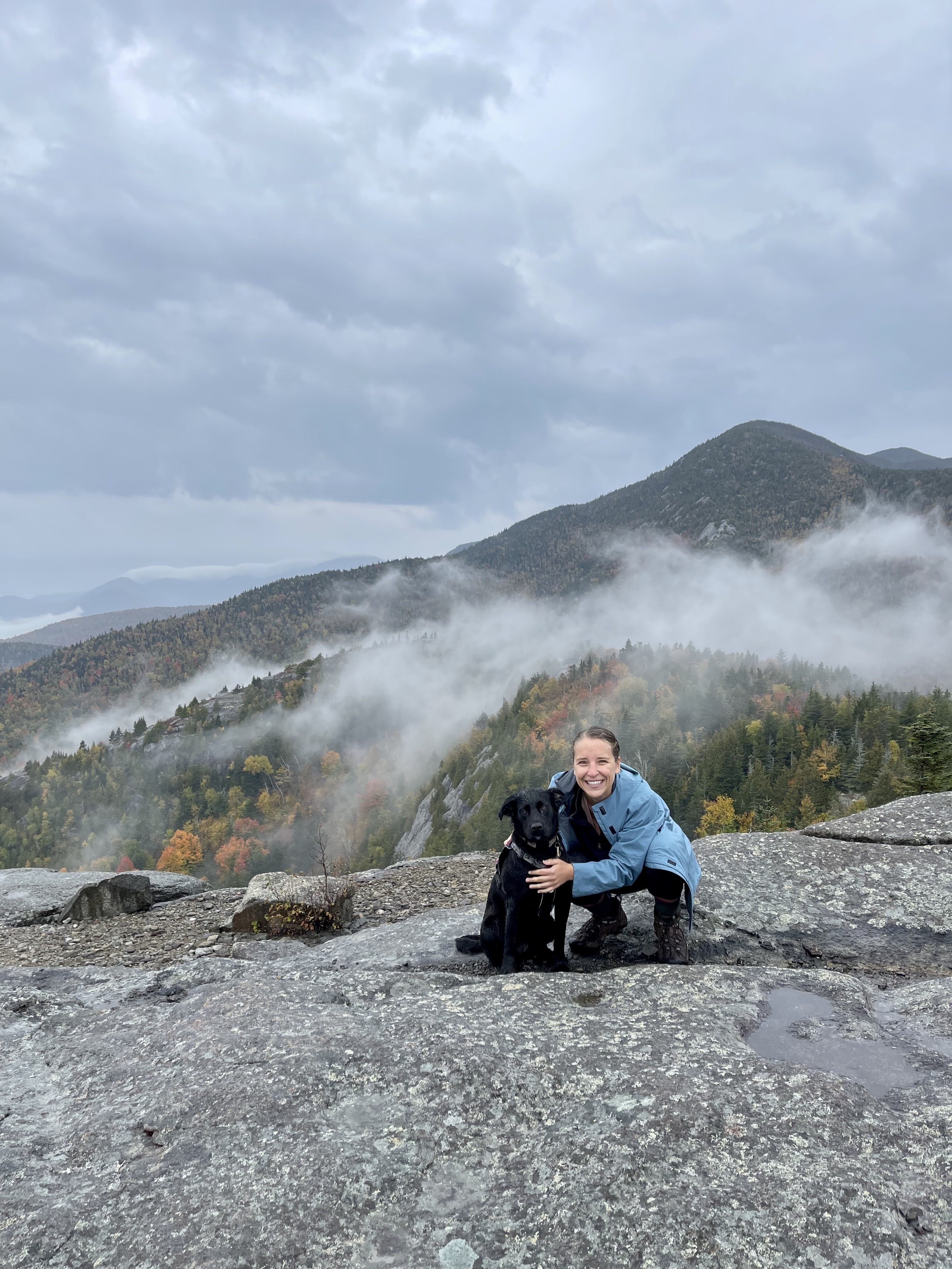 A smiling woman sits on an expanse of summit rock, hugging a black dog.