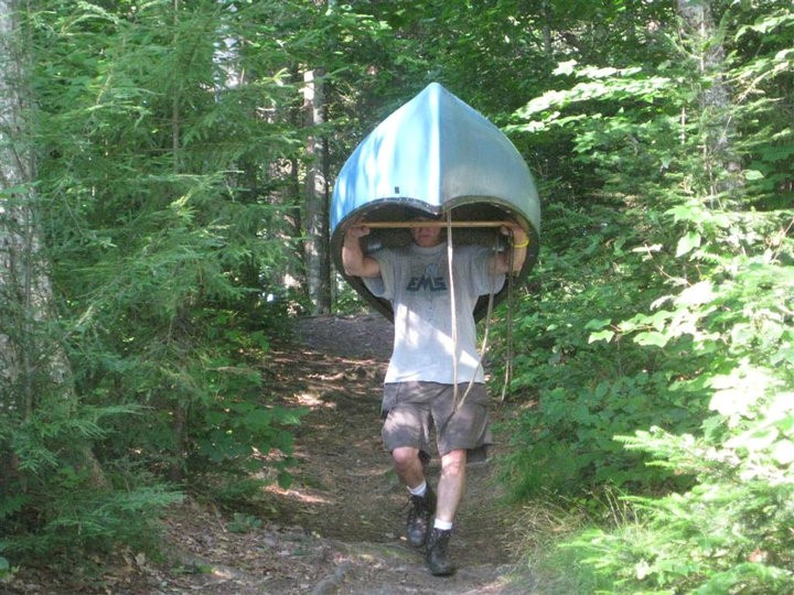 A man walks through the woods with a canoe held above his head.