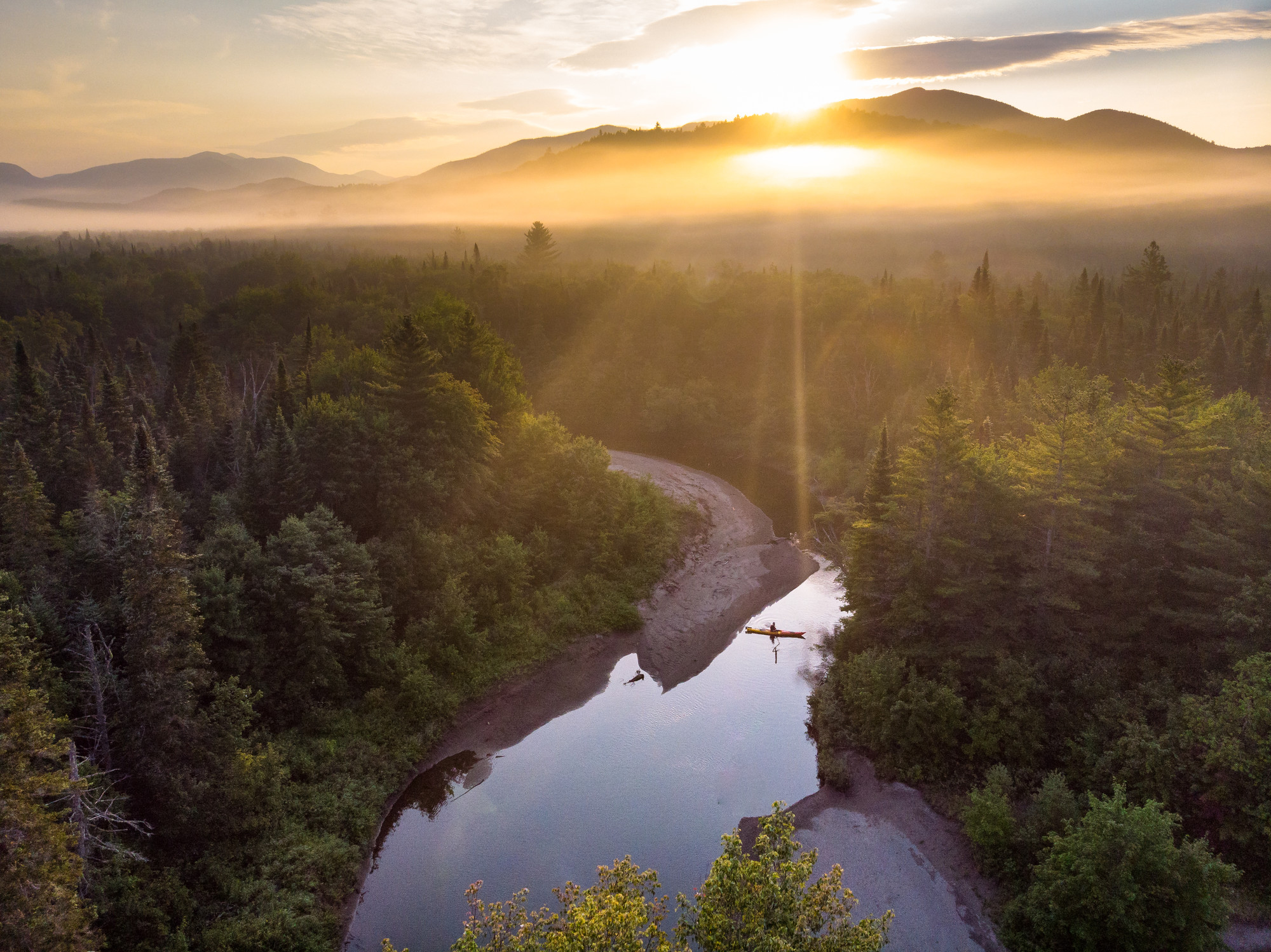 An aerial view of a kayak on a river surrounded by forest and mountains.