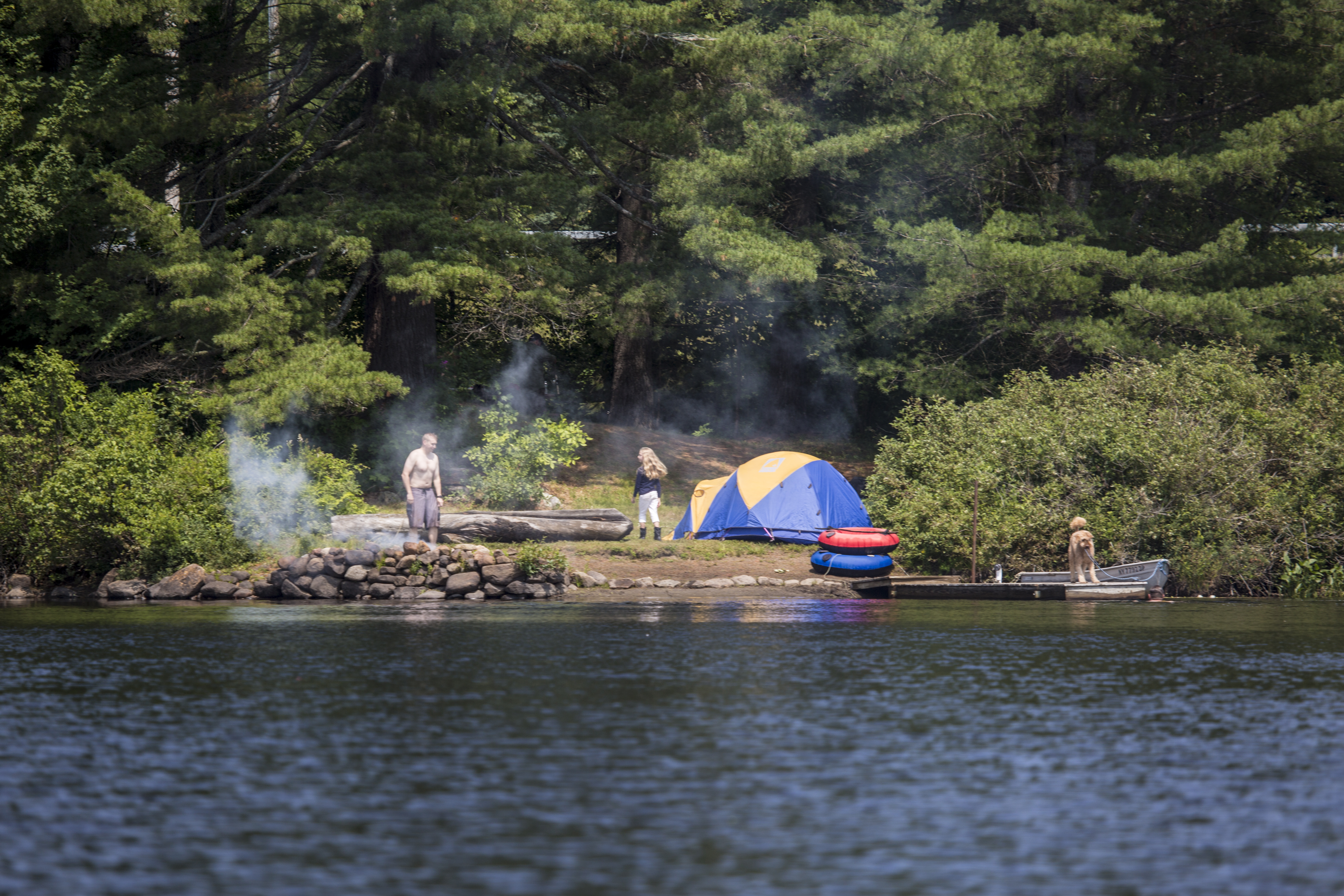 A photo shot from the water of two people and their dog camping on a rocky shore.