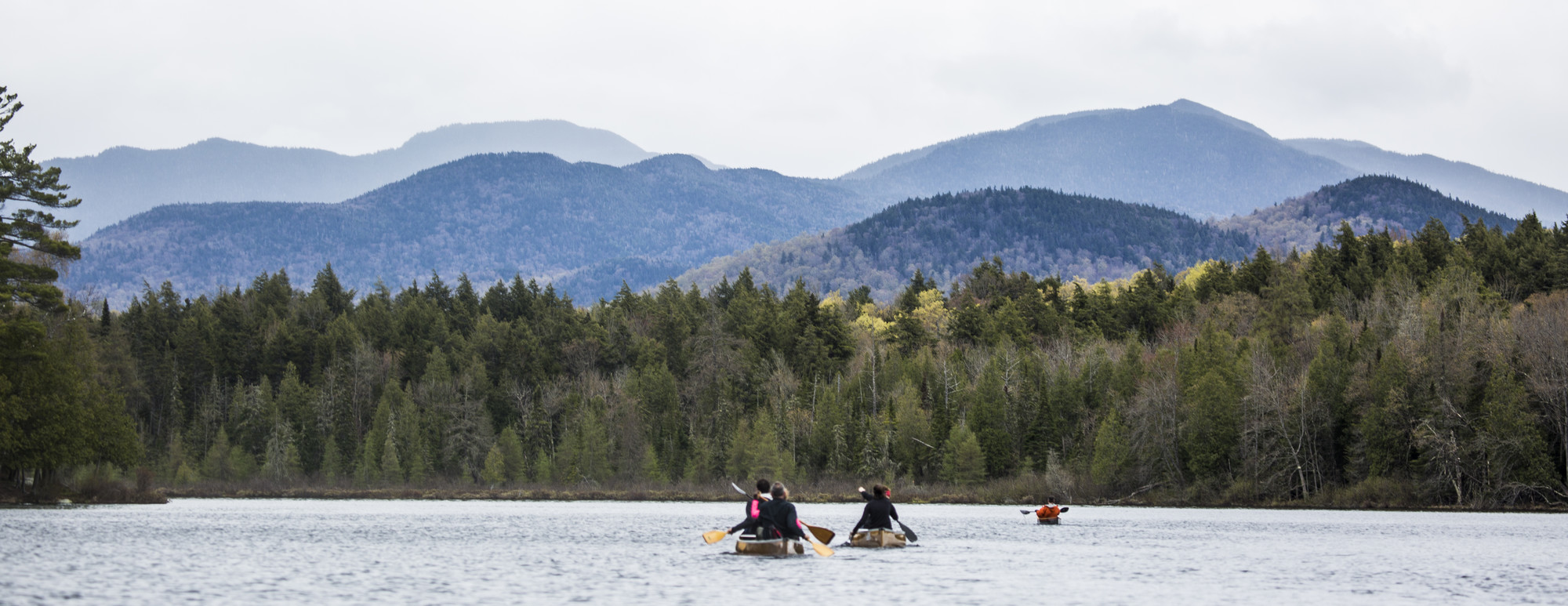 Two canoes and a kayak paddle away from the camera and towards the backdrop of the Adirondack mountains 