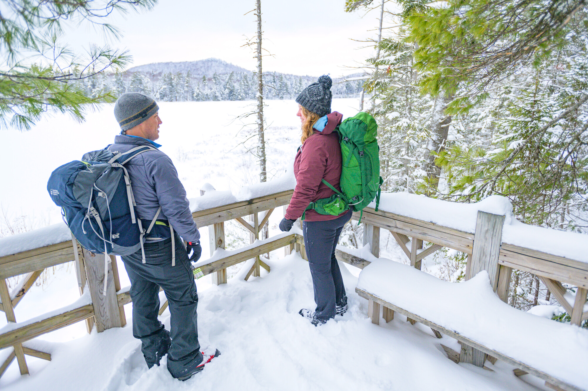 A man and woman look out on a frozen lake from a snowy lookout platform.