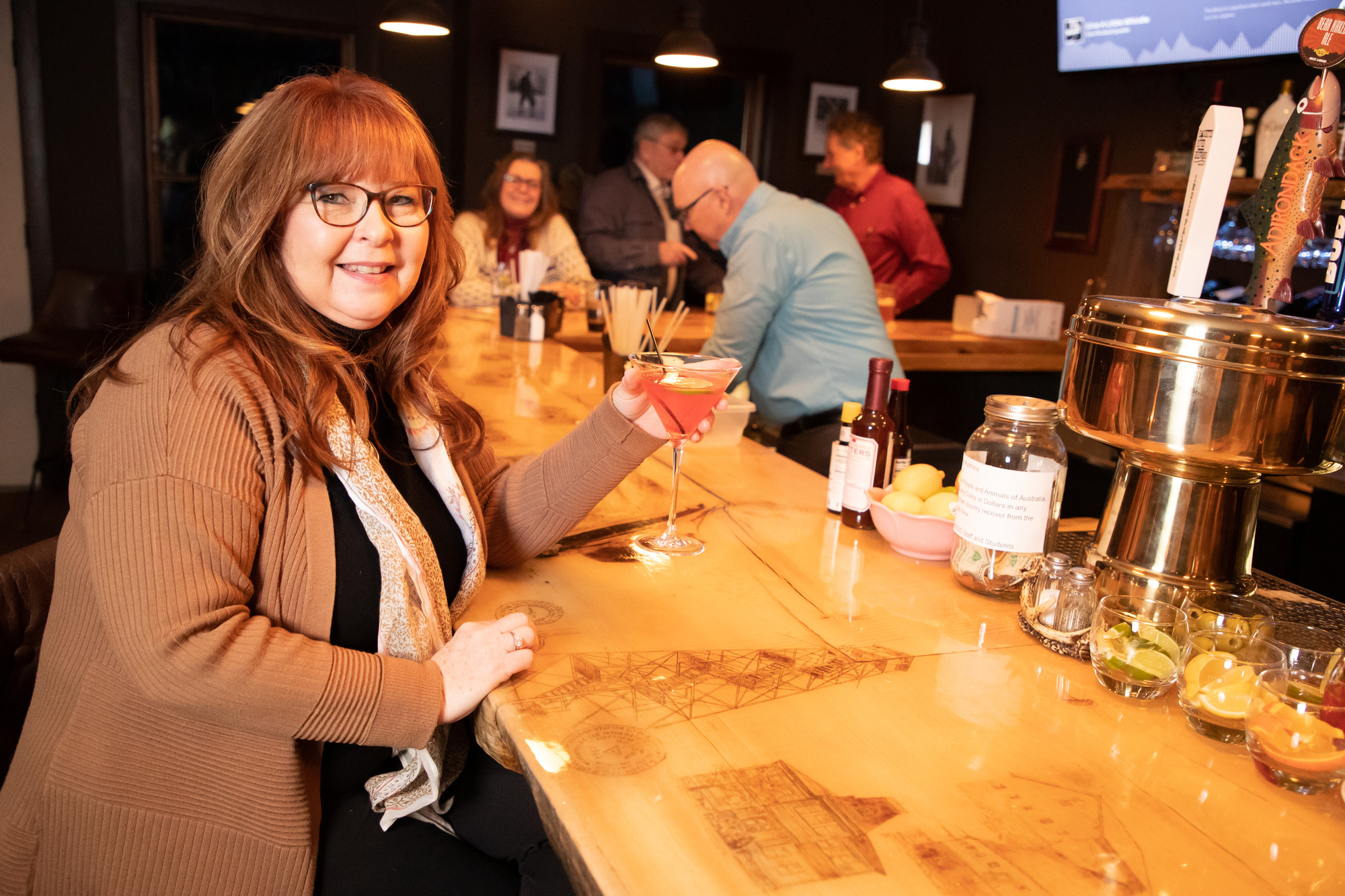 A red-haired woman wearing glasses holds a cocktail at a bar.