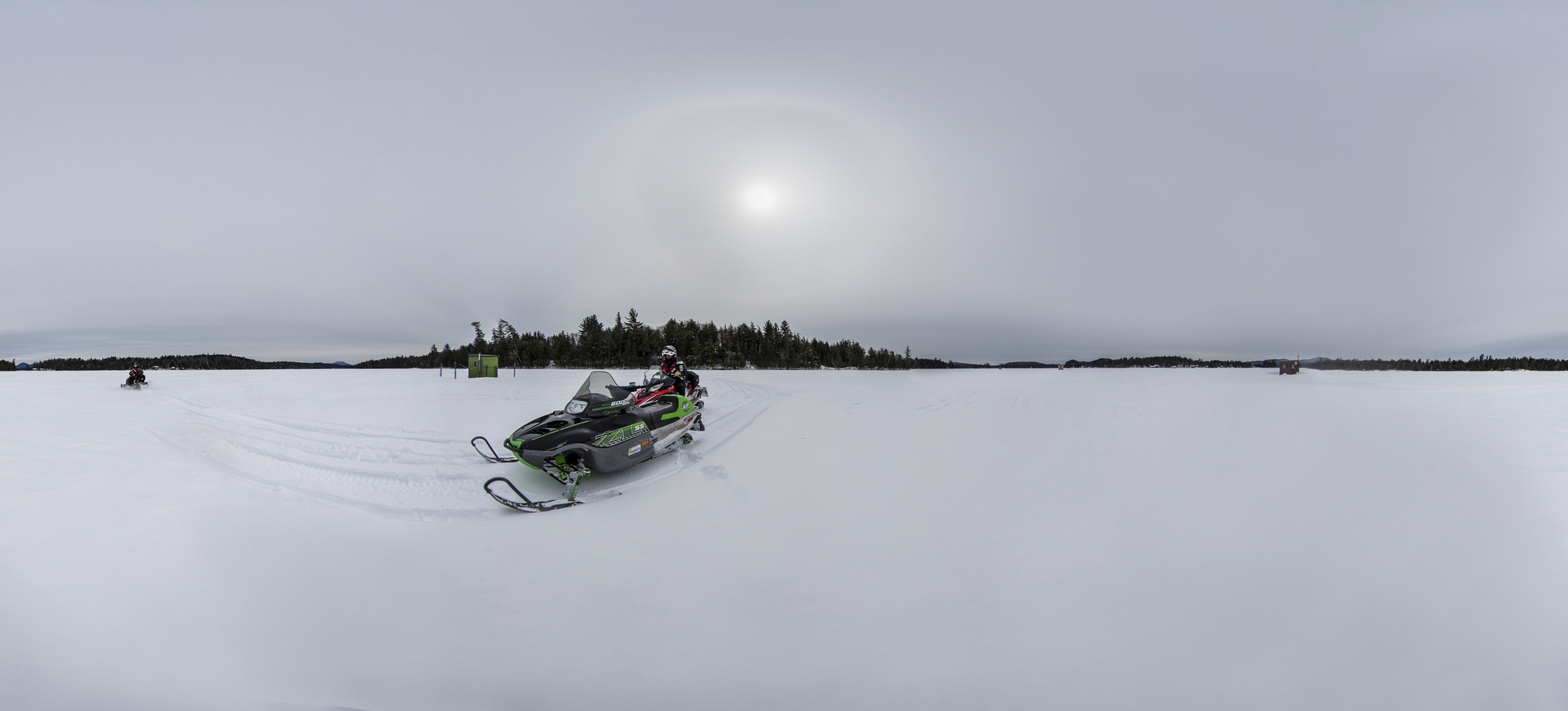 A snowmobiler drives a snowmobile across a large frozen Adirondack lake with a fishing ice shanty in the distance.