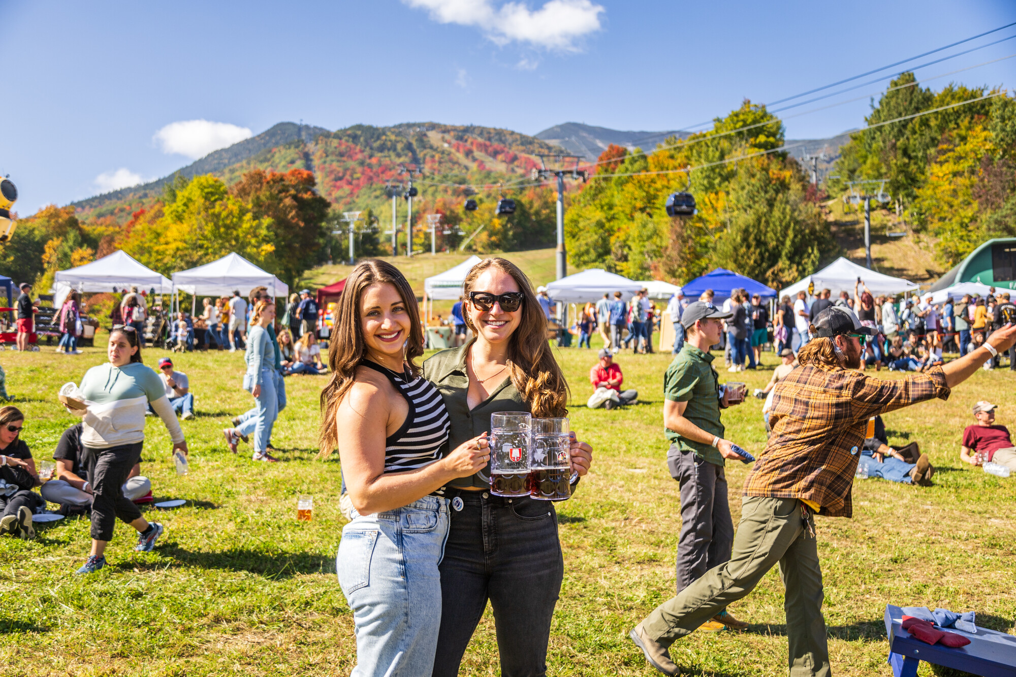 Two women smile for the camera while a fall festival, with tents and crowds in the background.
