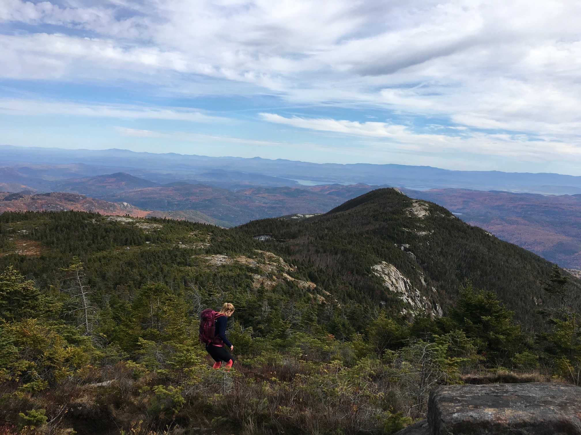 A blonde woman descends a mountain trail surrounded by low, Alpine vegetation.