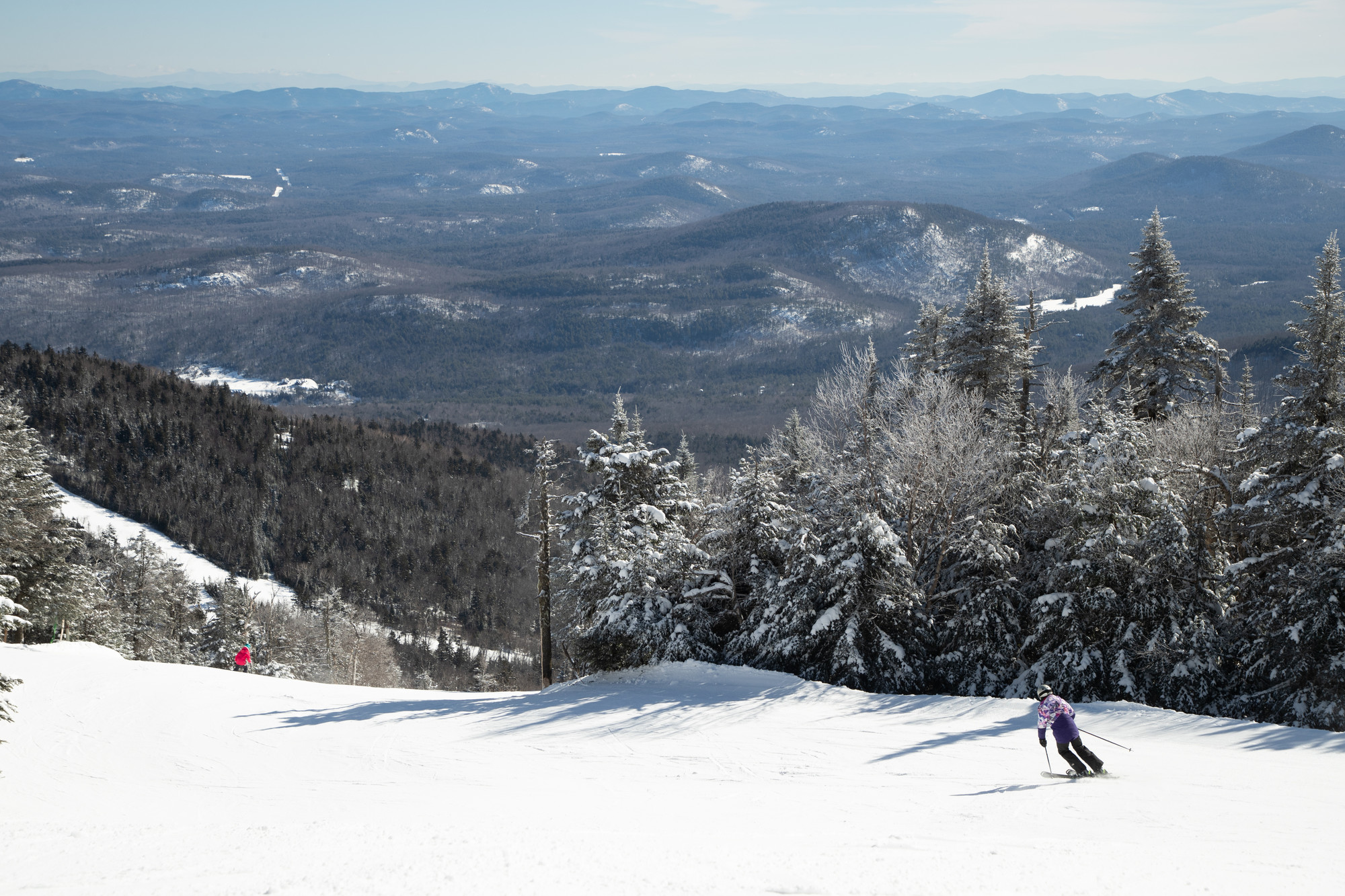 A lone skier descends a snowy slope with a sea of mountain peaks in the distance