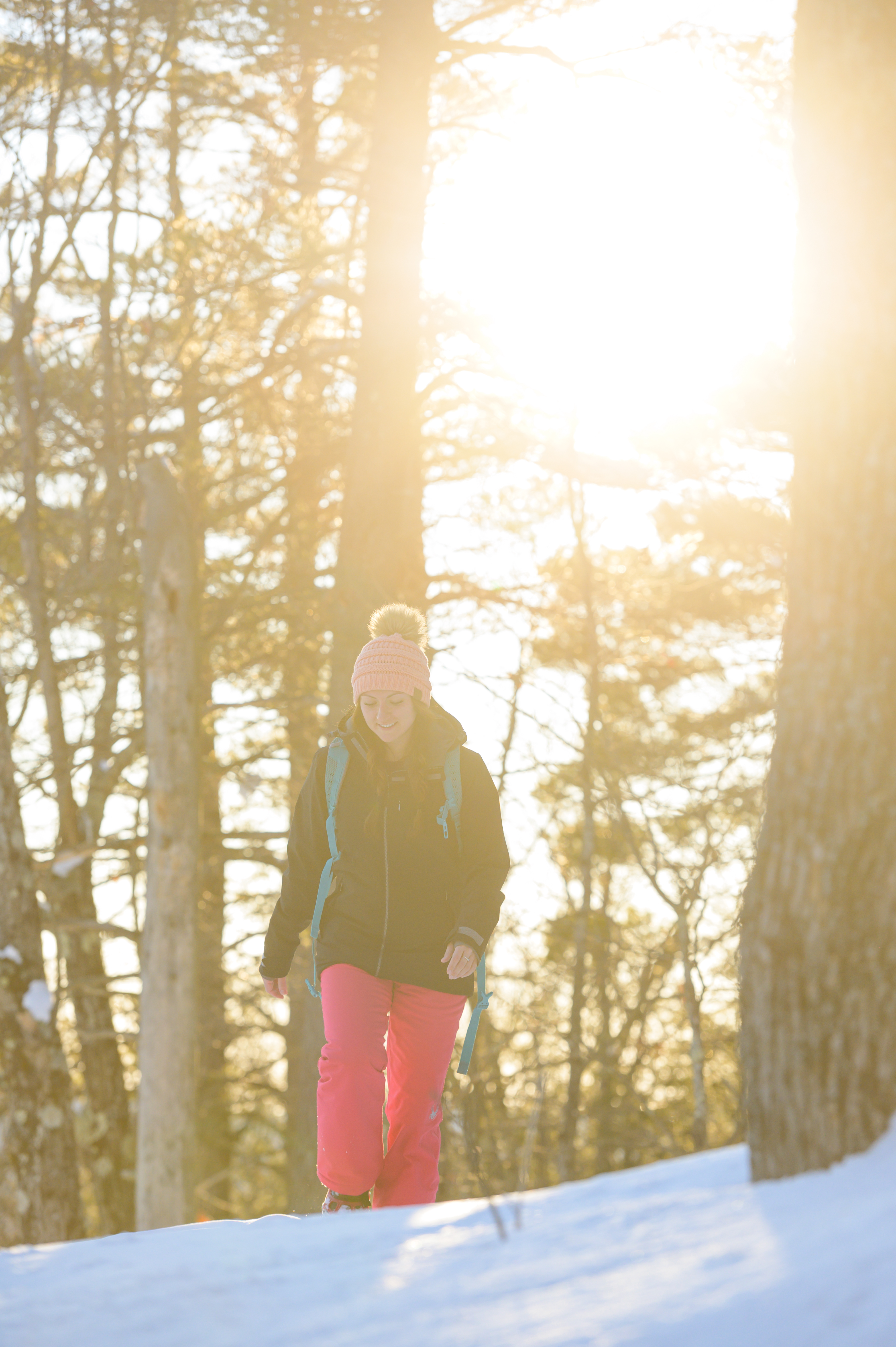 Woman walking on a snowy trail with sun shining through the trees.