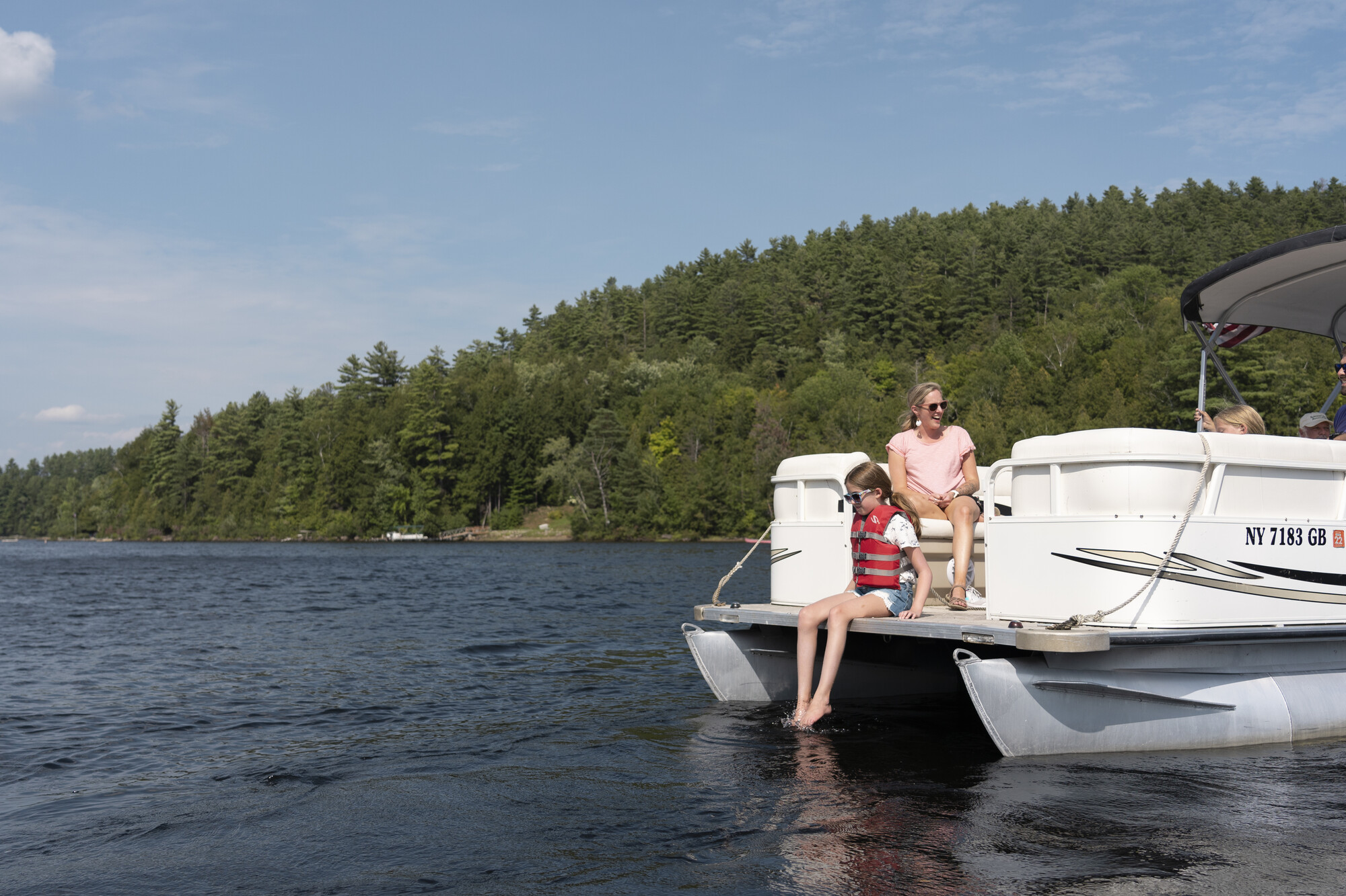 A woman sits on the end of a boat near a child in a life jacket who is about to go swimming.