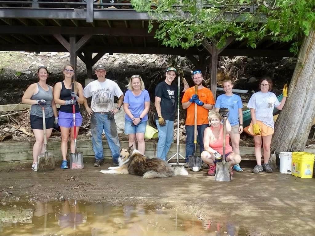 A group of adults in summer clothing, holding shovels, pose on a dirt-covered shoreline.