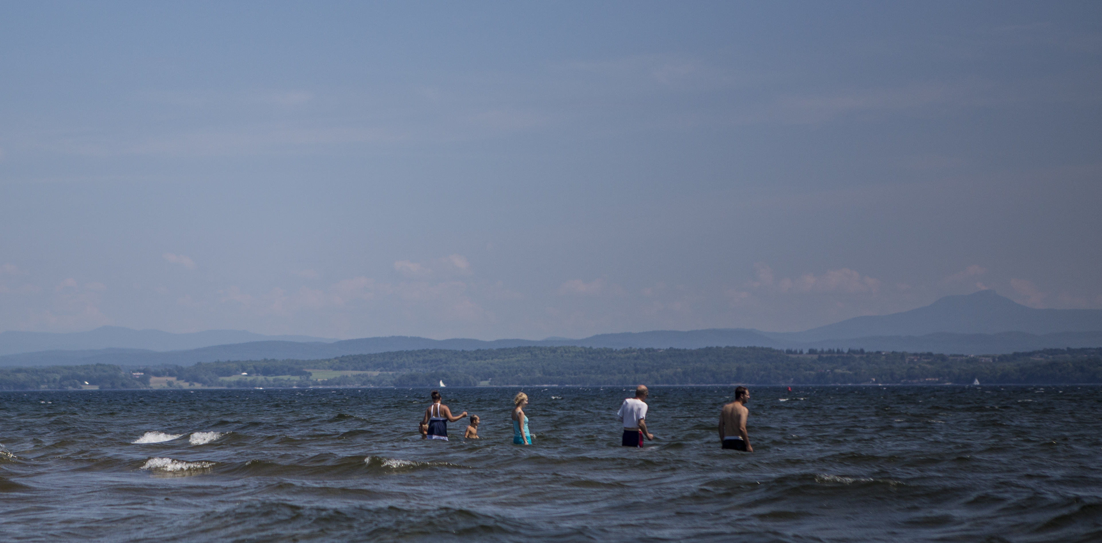 Adults and children play in the waters of Lake Champlain with the Green Mountains beyond.