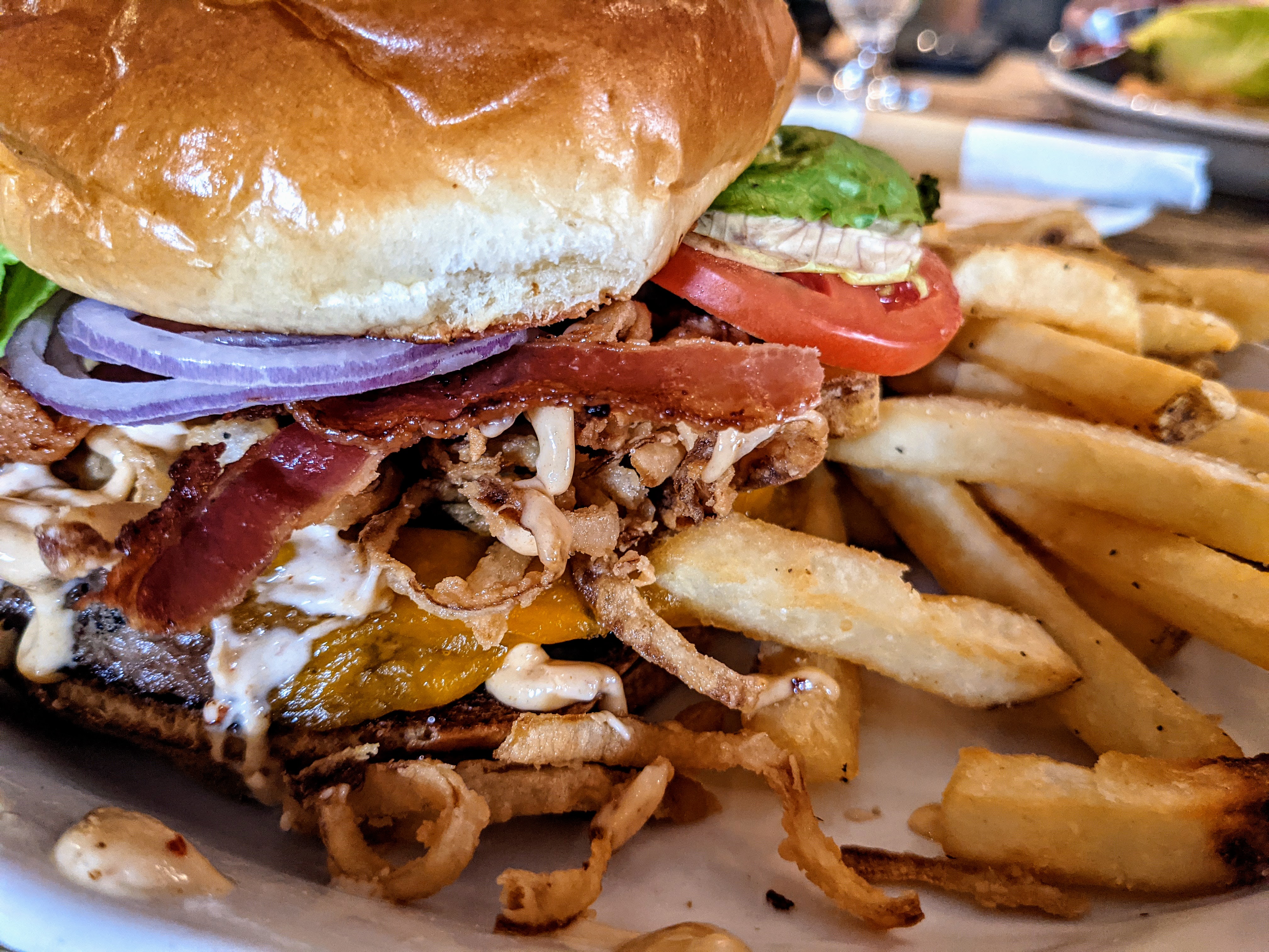 A close-up image of a burger topped with lettuce, tomato, onion, and bacon with a crispy side of fries