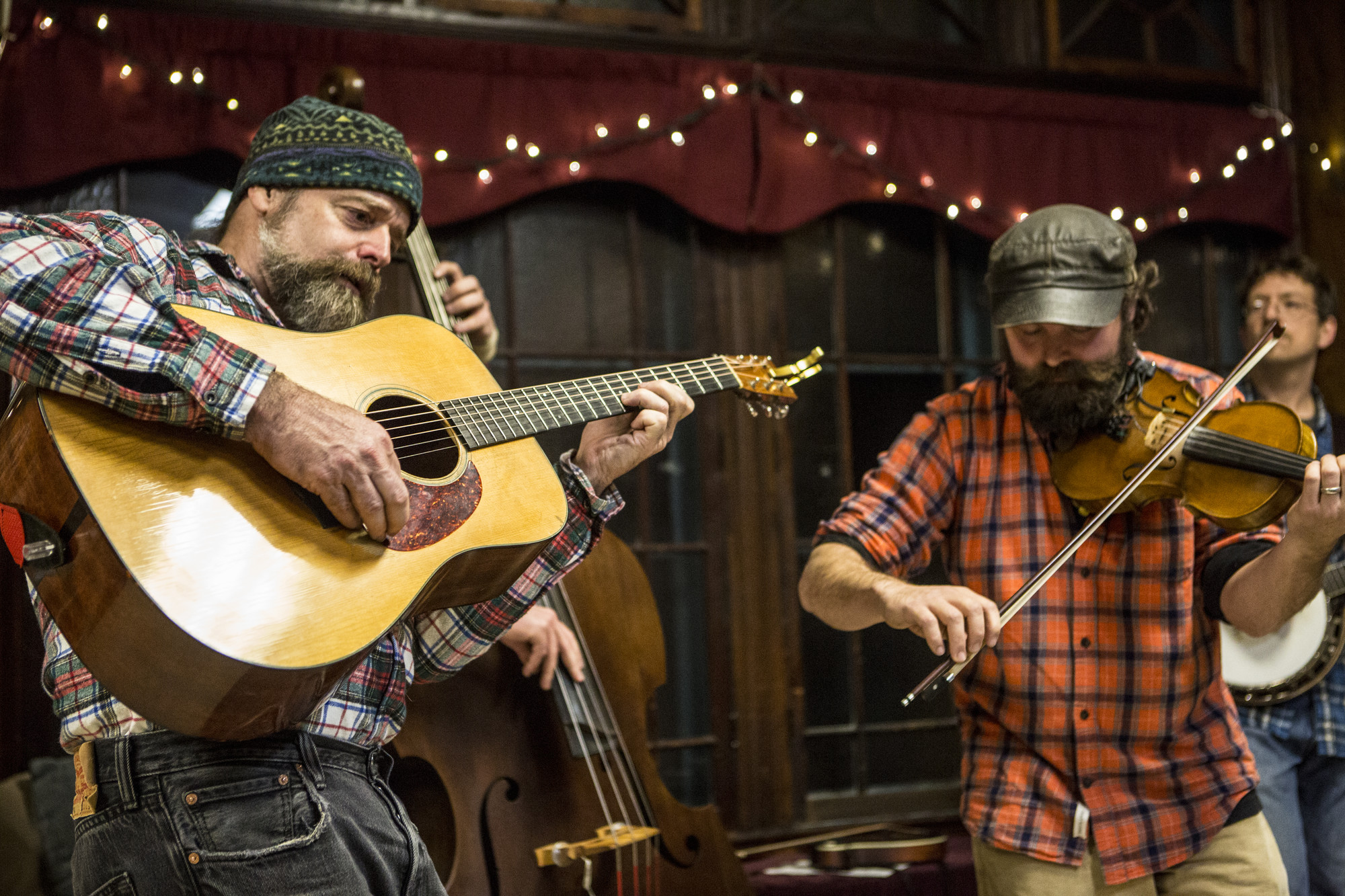 A folk group in plaid performs on guitar, bass, and fiddle at an indoor music event.