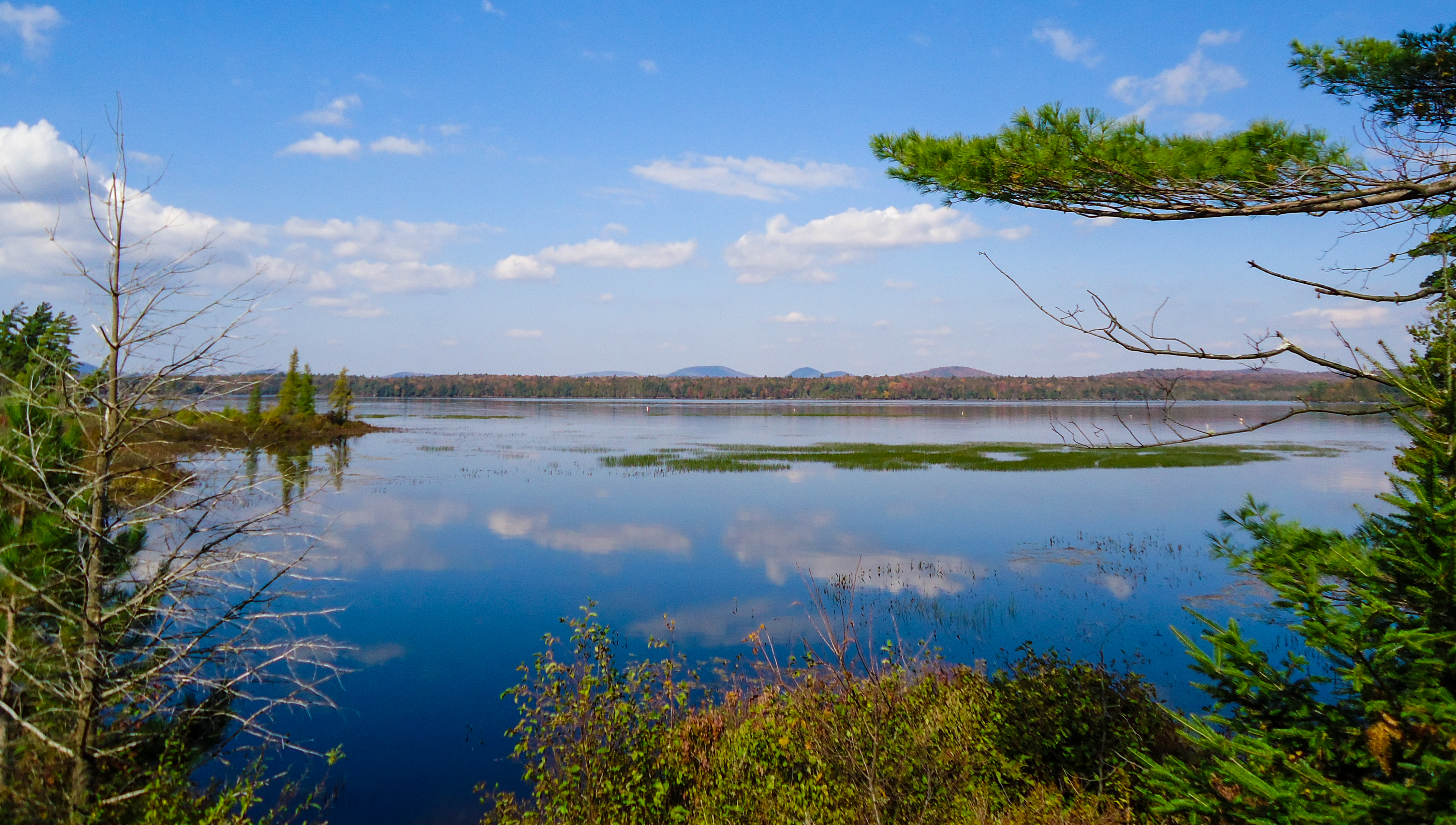 A placid blue lake with clouds reflected and foliage colors and mountains in the background.